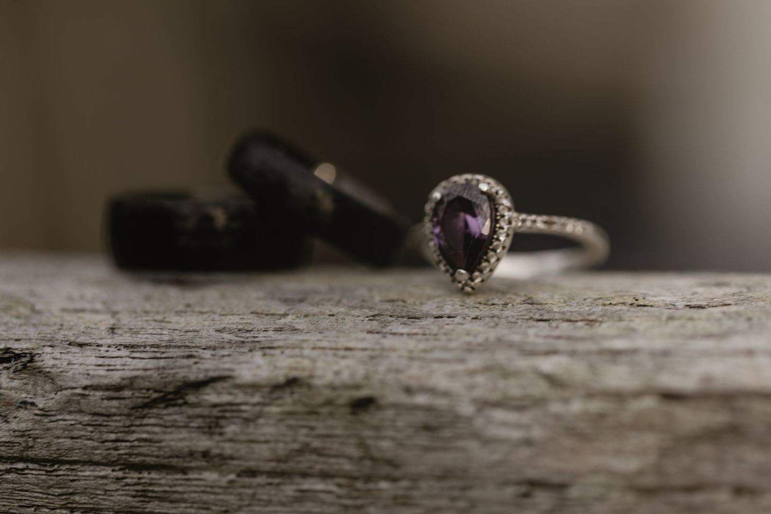 A delicate vintage-style ring with a purple gemstone and diamond halo sits on weathered wood surface.