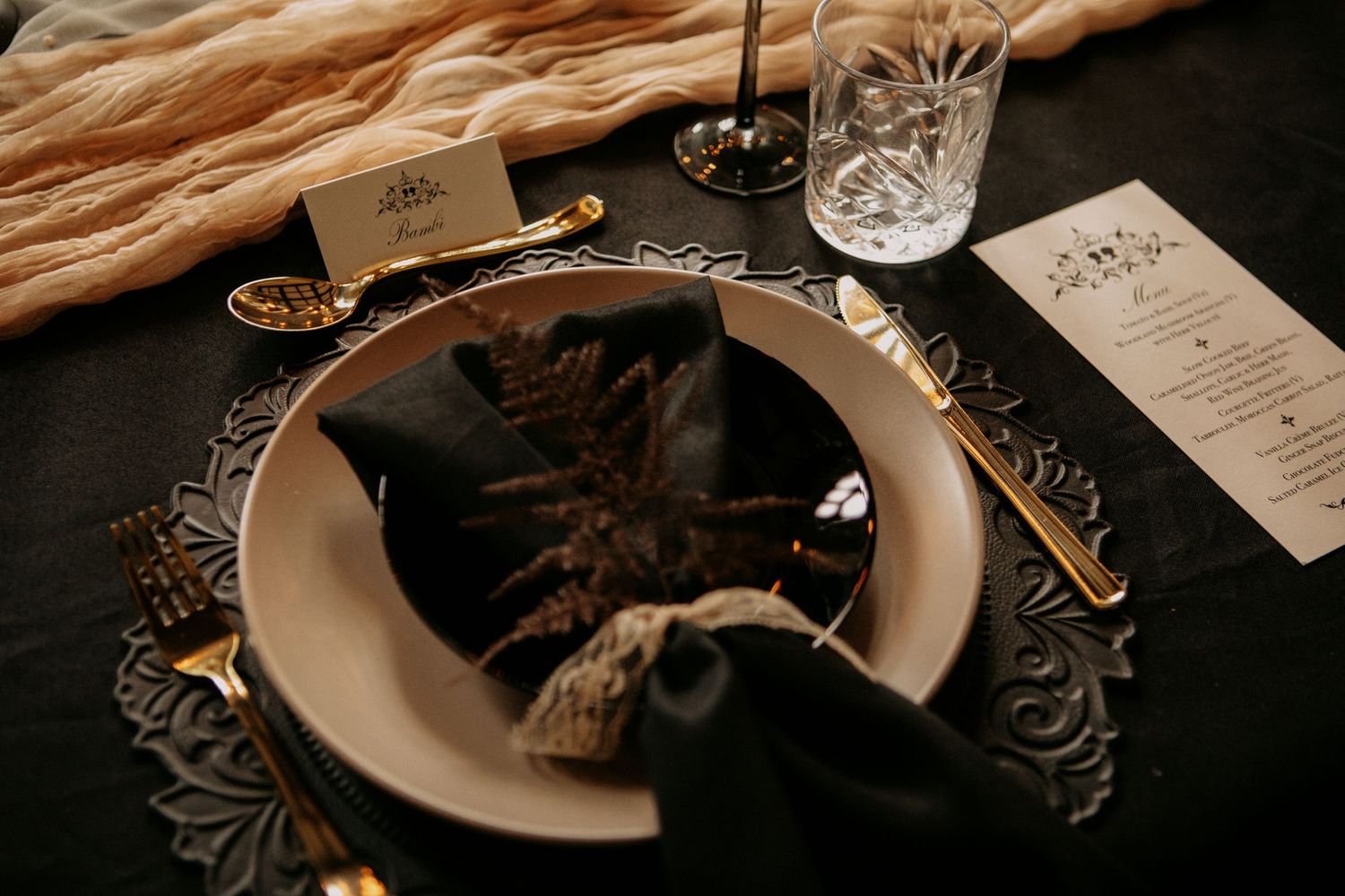 Elegant table setting with ornate silver charger plate, black napkin, menu card and glassware on dark background.