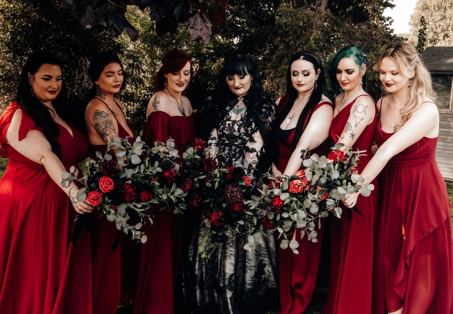 Wedding party in red bridesmaid dresses and a gothic bride in black pose with dramatic burgundy floral bouquets.