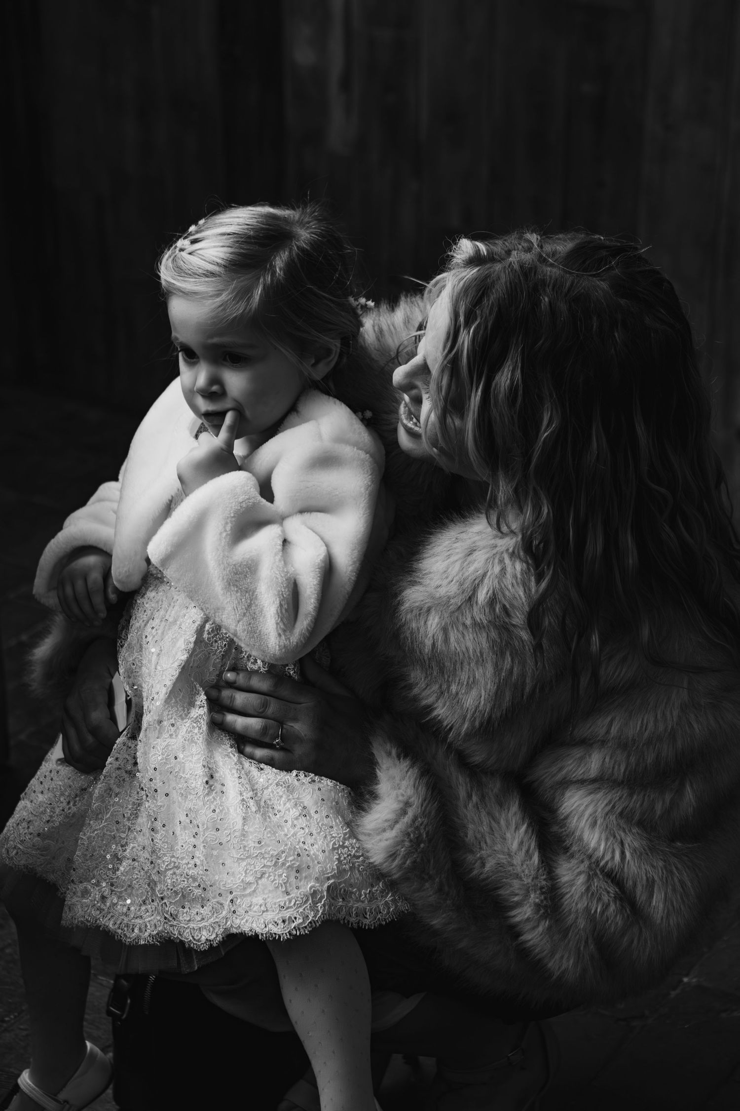 Black and white portrait of tender moment with child in white dress and person in fur coat.