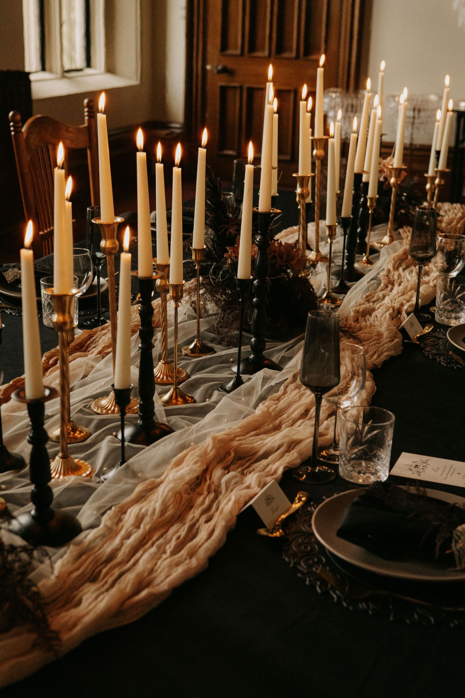 Gothic table setting with black candlesticks, white taper candles, and ethereal draped fabric on dark tablecloth.