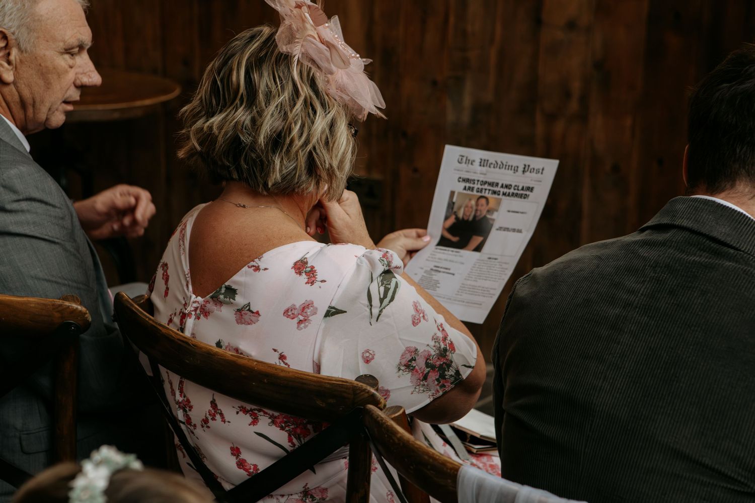 Woman in floral dress reads a newspaper while seated between two people in formal attire.