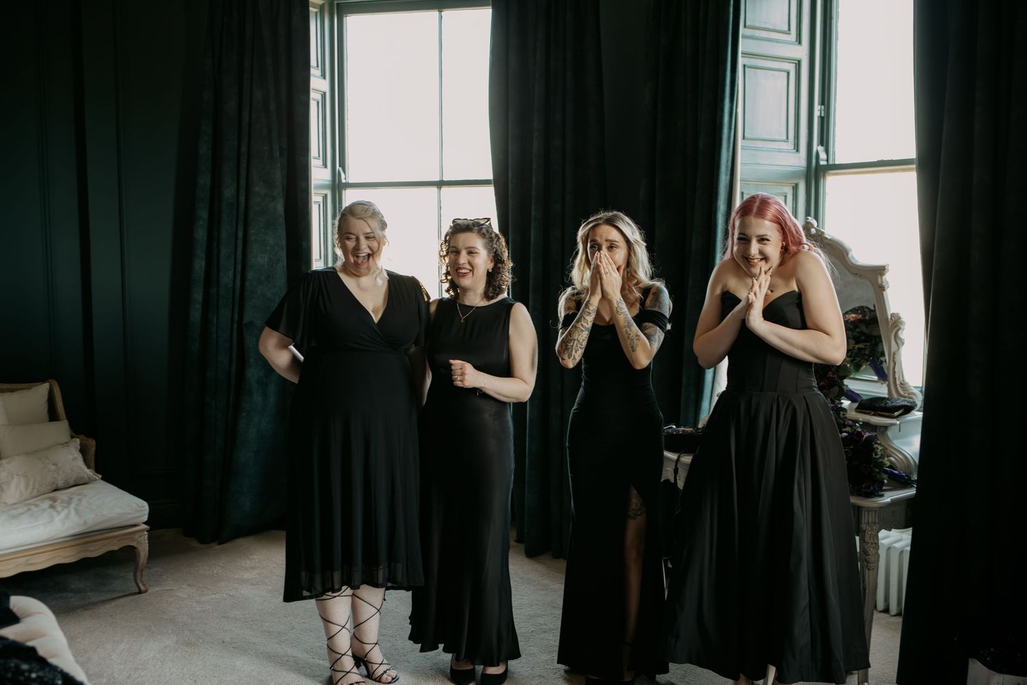 Four women wearing elegant black dresses pose together in front of a bright window in a stylish interior space.