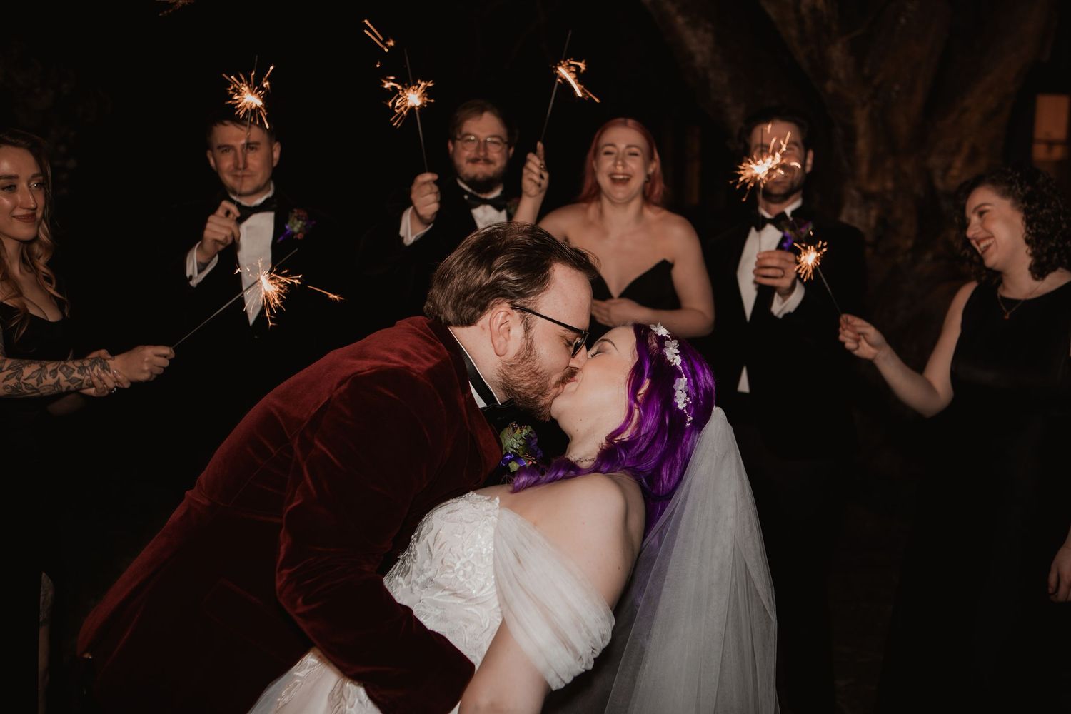 A couple shares a dramatic dip kiss at their wedding reception while guests celebrate with sparklers in the background.