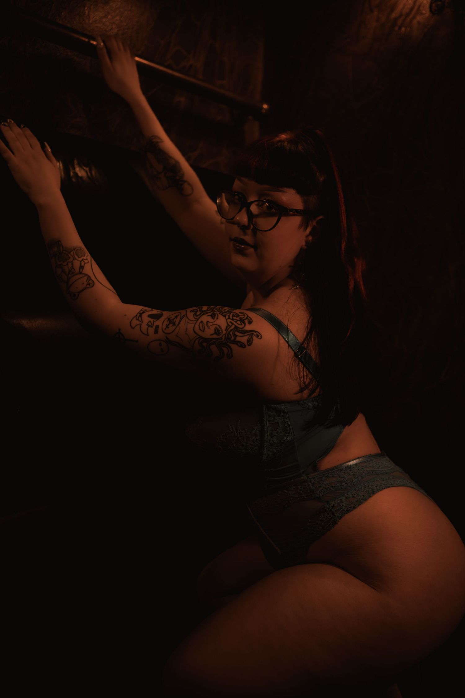 A moody boudoir photo shows a woman in lingerie with tattoos posing dramatically in dark lighting.