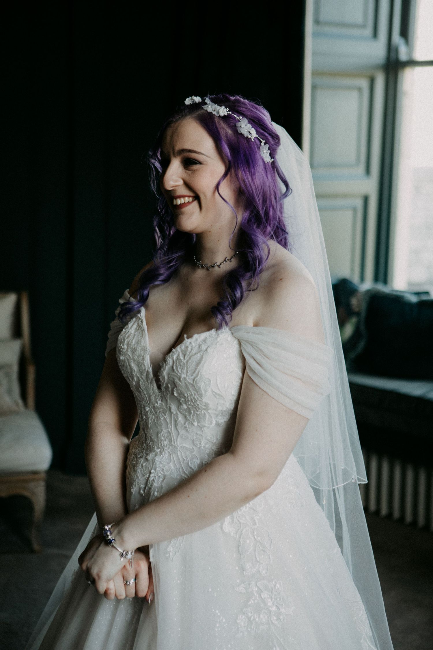 Joyful bride in lace wedding dress and flowing veil laughs while showing off purple hair and delicate floral headpiece.