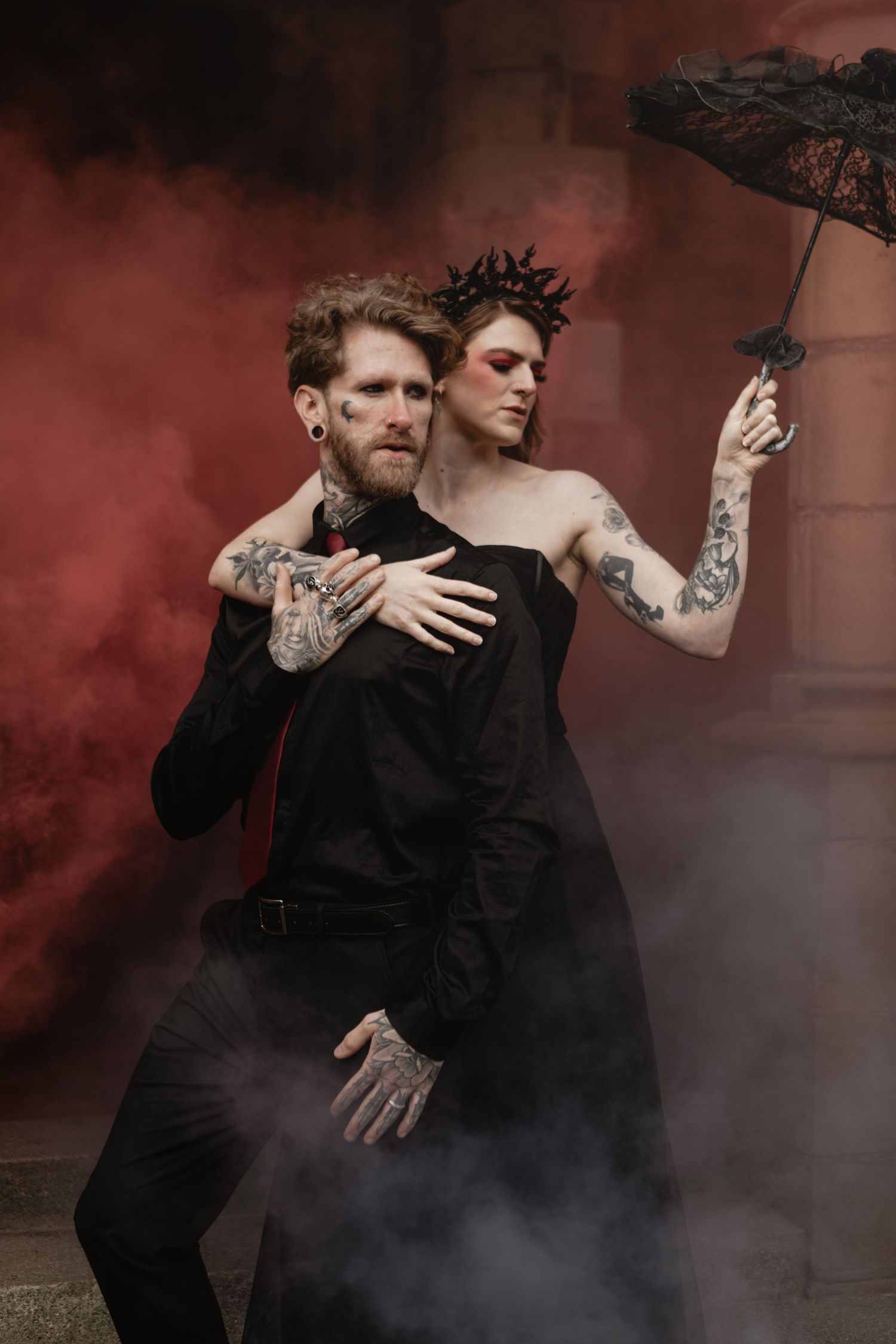 Two figures in black gothic attire pose dramatically with umbrella against moody red smoke-filled backdrop.