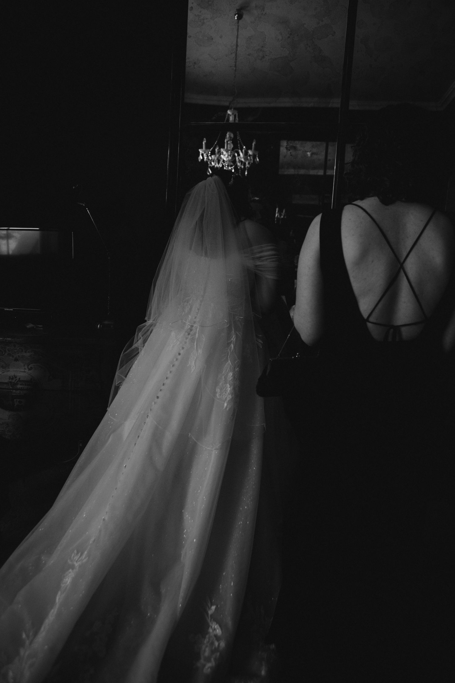 Black and white photo showing the back of a long, flowing wedding dress veil in a dimly lit room with a chandelier.