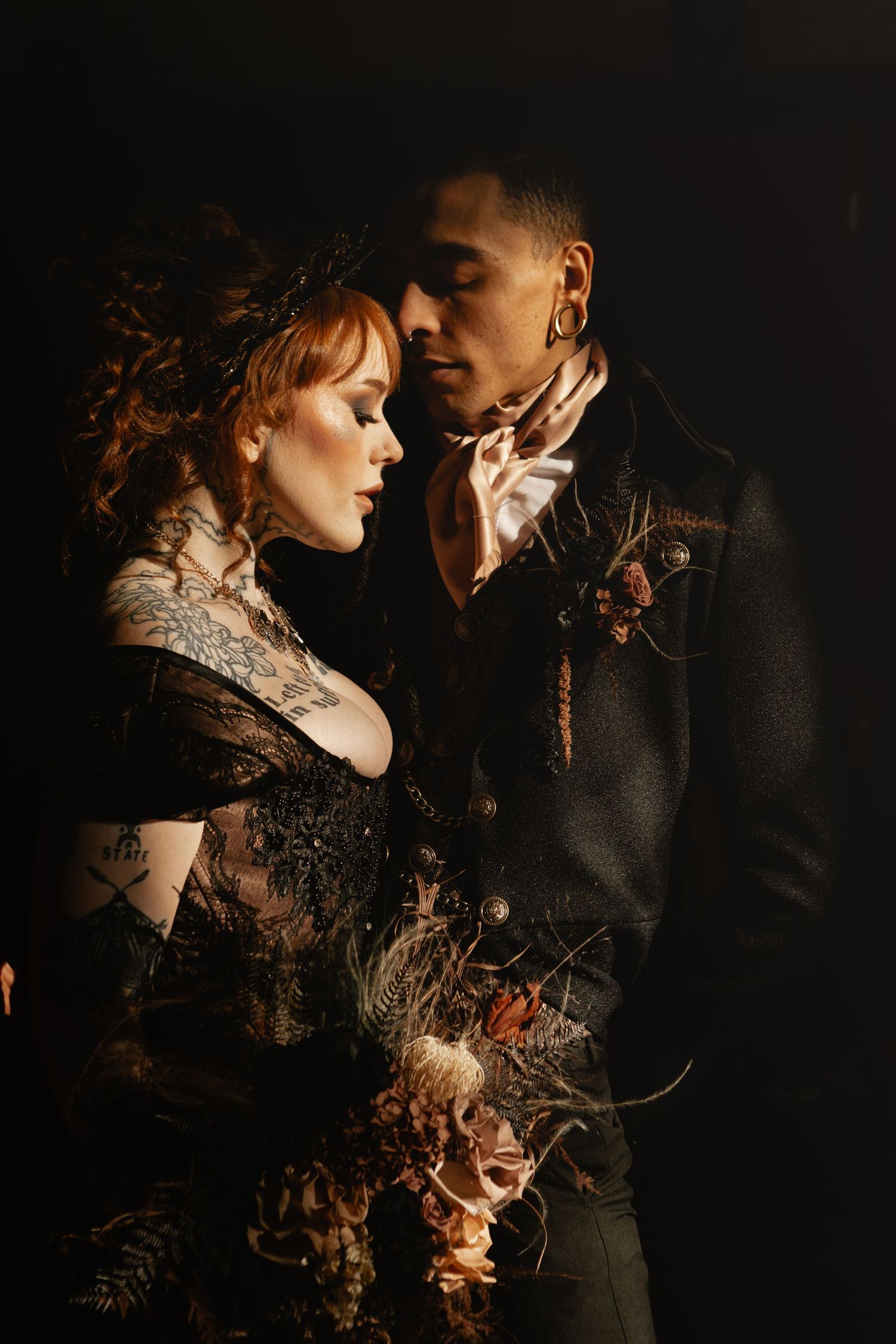 Romantic gothic-style portrait of a couple in dark vintage clothing with moody lighting and dried floral elements.