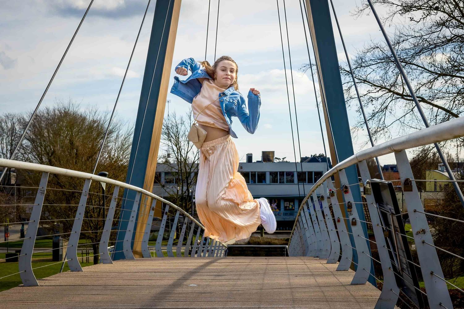 A joyful person wearing a beige dress jumps with blue balloons on a modern bridge with metal railings.