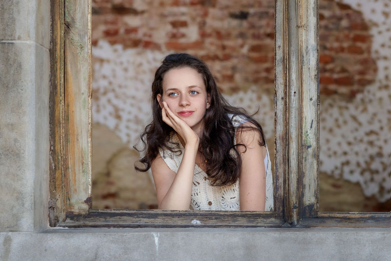 A person rests their chin on their hand while gazing through an old rustic window frame with exposed brick behind.