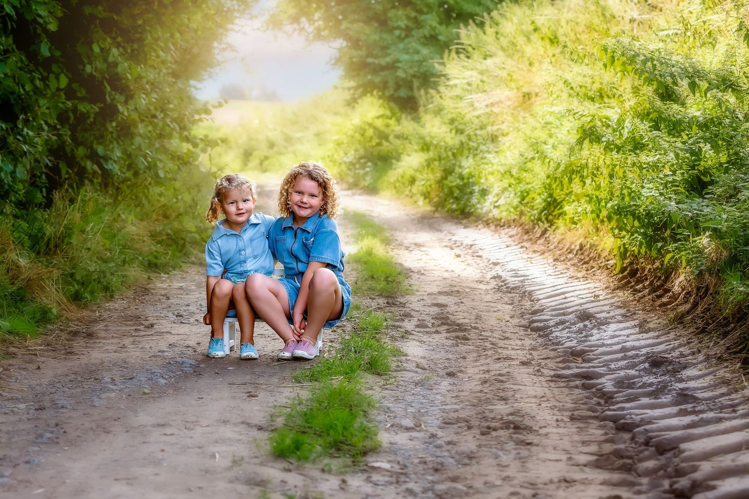 Two figures in matching blue outfits sit together and smile on a sunny dirt path surrounded by greenery.