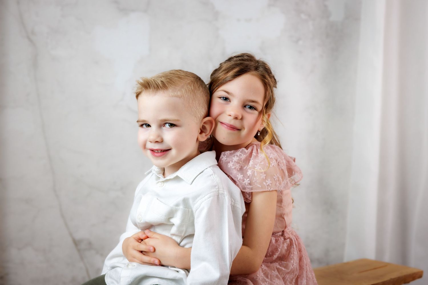Two children pose together against a gray backdrop, one wearing white and one in a pink dress.