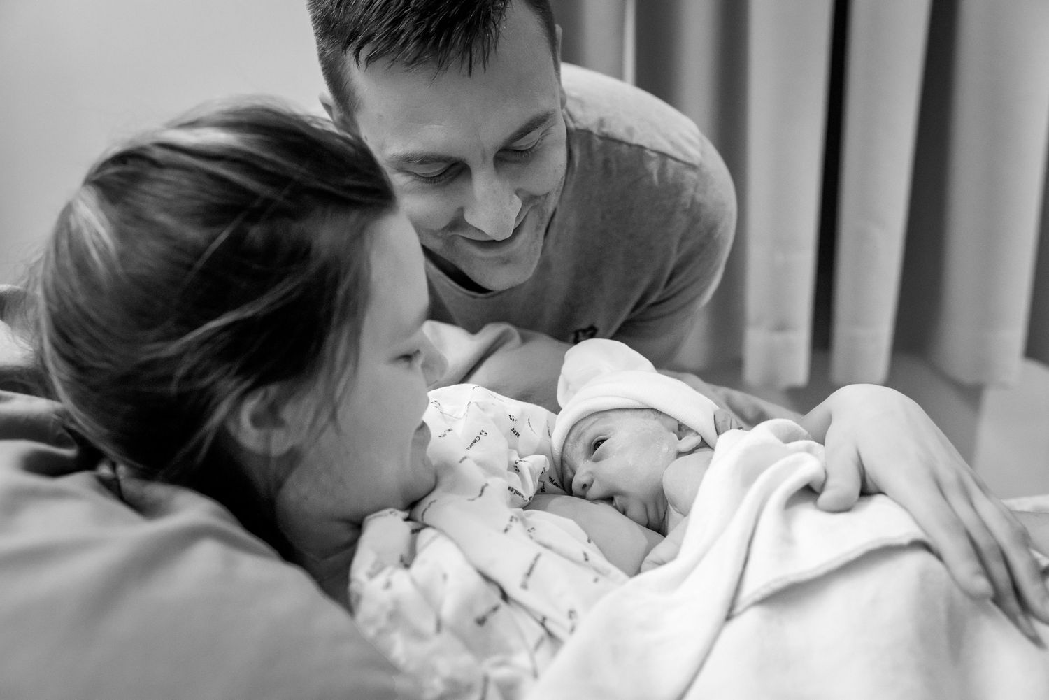 Black and white photo of parents sharing an intimate moment with their newborn baby in a hospital.