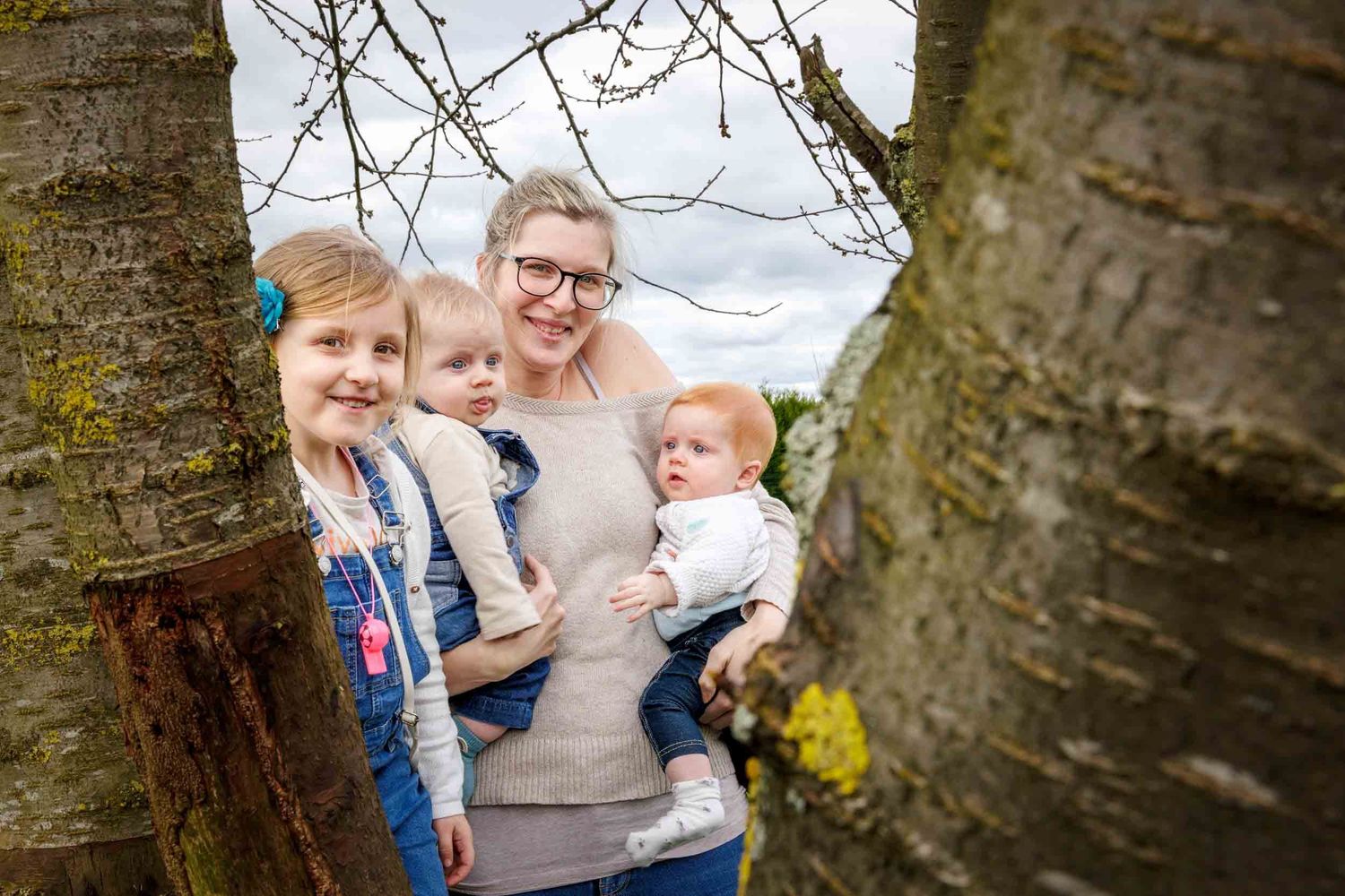 A family poses between tree trunks on a cloudy day in casual outdoor attire.