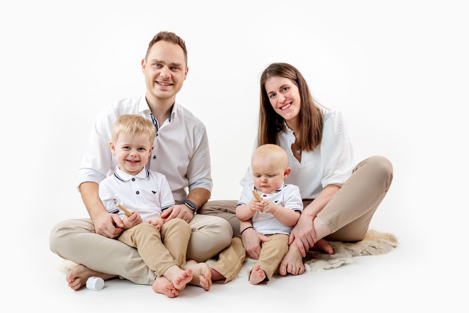 Family sits together on white background wearing neutral colors and matching casual outfits.