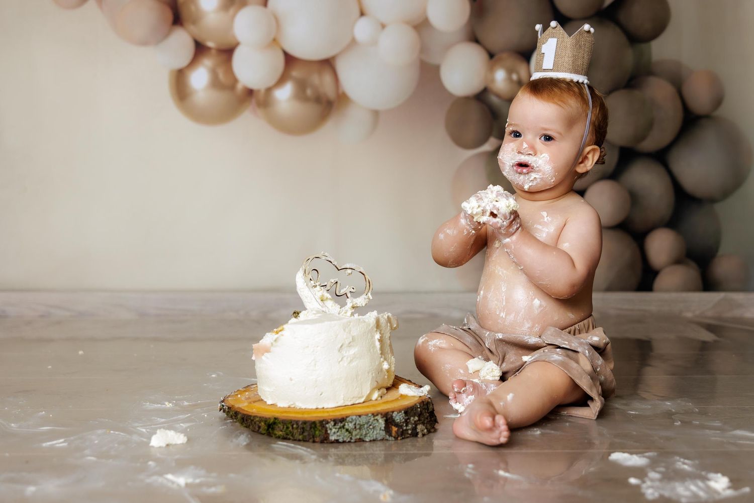 A cake smash photo session with white and gold balloon decorations, featuring a small cake on a wooden slice.