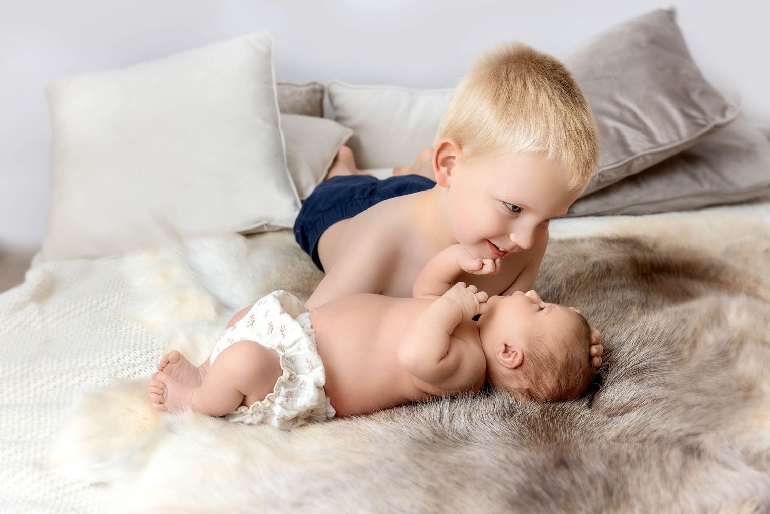 Toddler wearing navy shorts lying on fluffy white blanket with newborn sibling among scattered pillows.