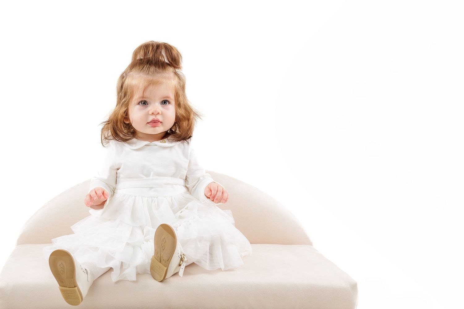 A child wearing a white lace dress sits on a cream-colored sofa with light brown hair styled up.