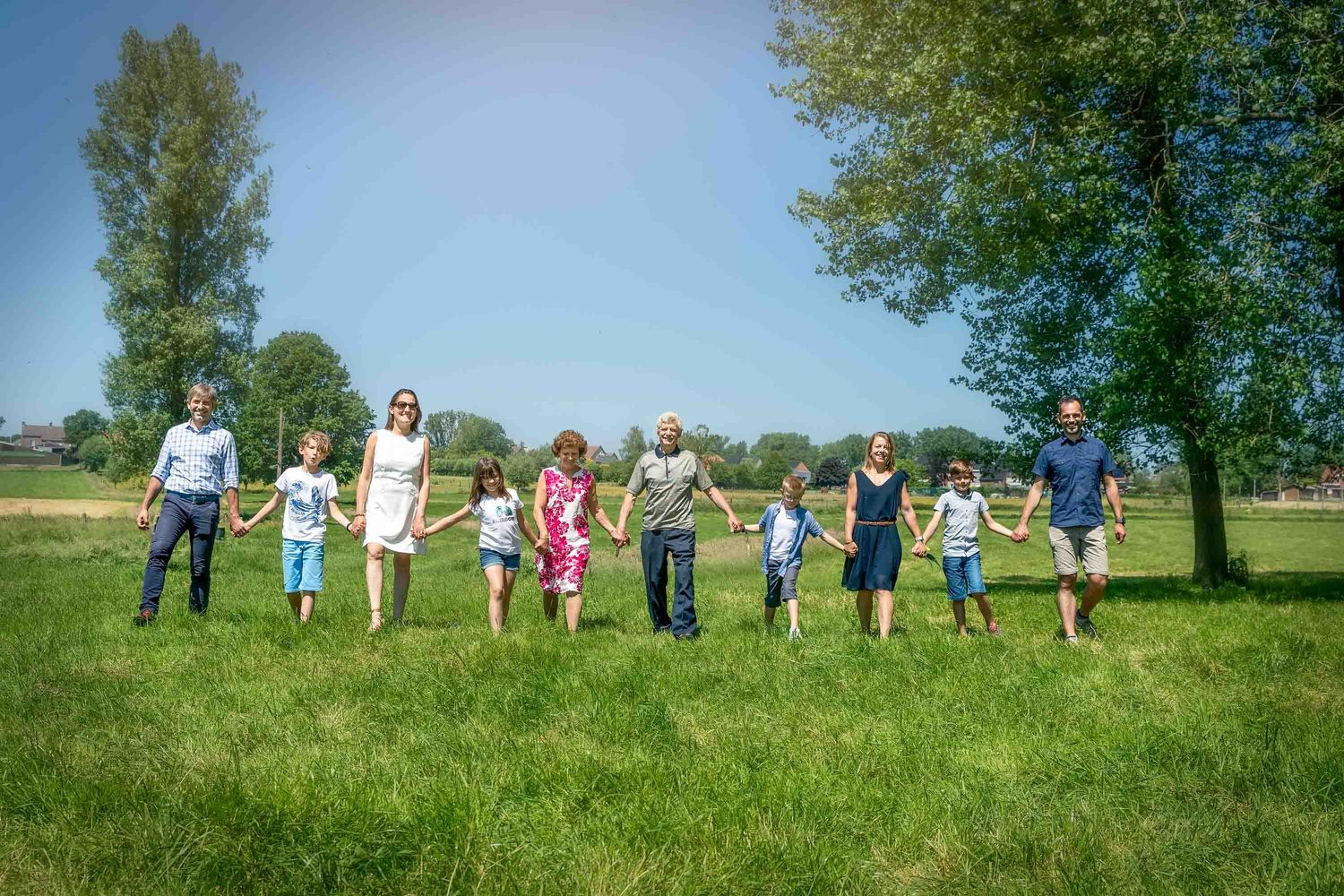 Group of people jumping together in a line on a grassy field on a sunny summer day with trees in the background.