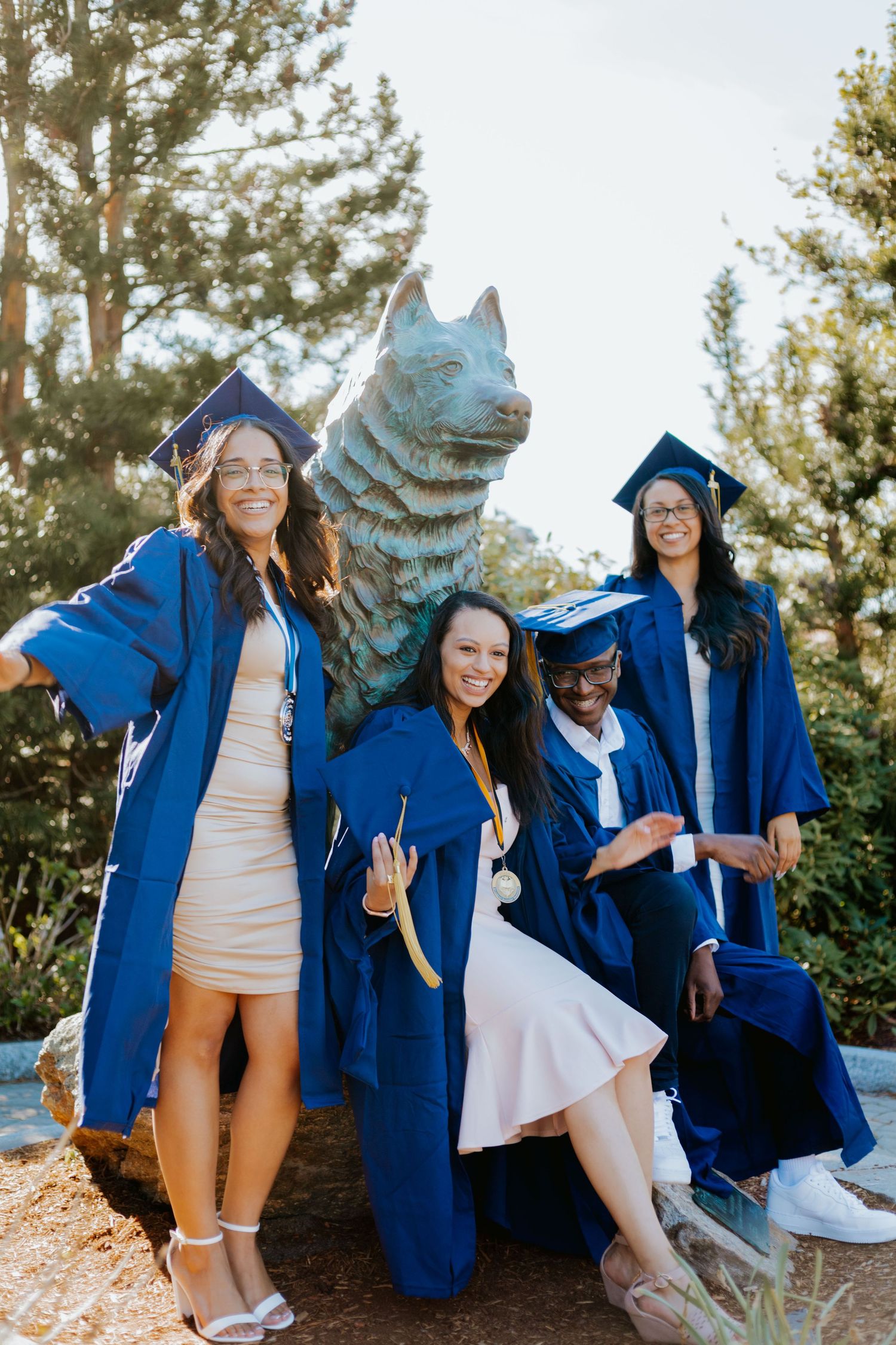 Graduates in blue robes posing with a large metallic wolf statue during commencement celebrations.