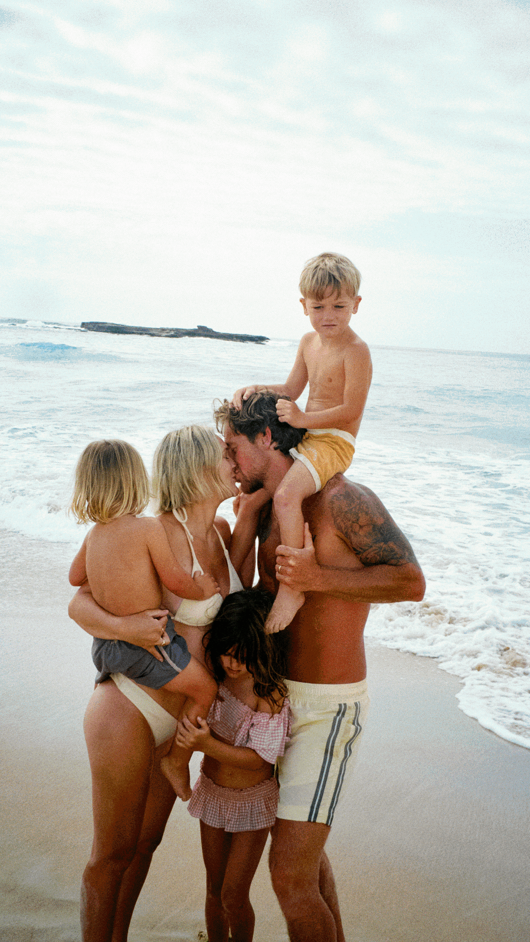 A loving family enjoys a sunny day together at the beach with ocean waves and sand in the background.