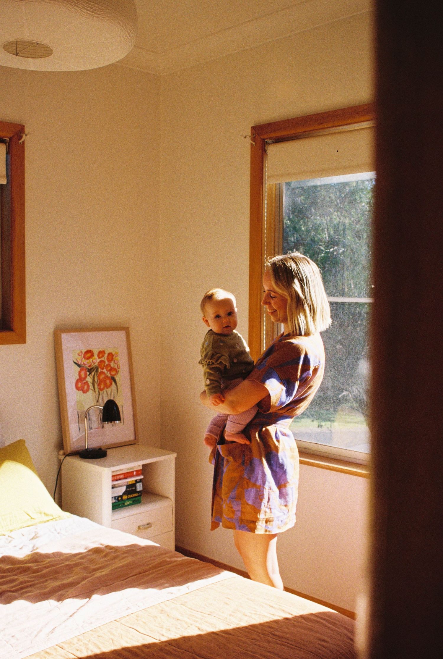 Sunlit bedroom interior with window, unmade bed, floral artwork, and figures standing in warm natural light.