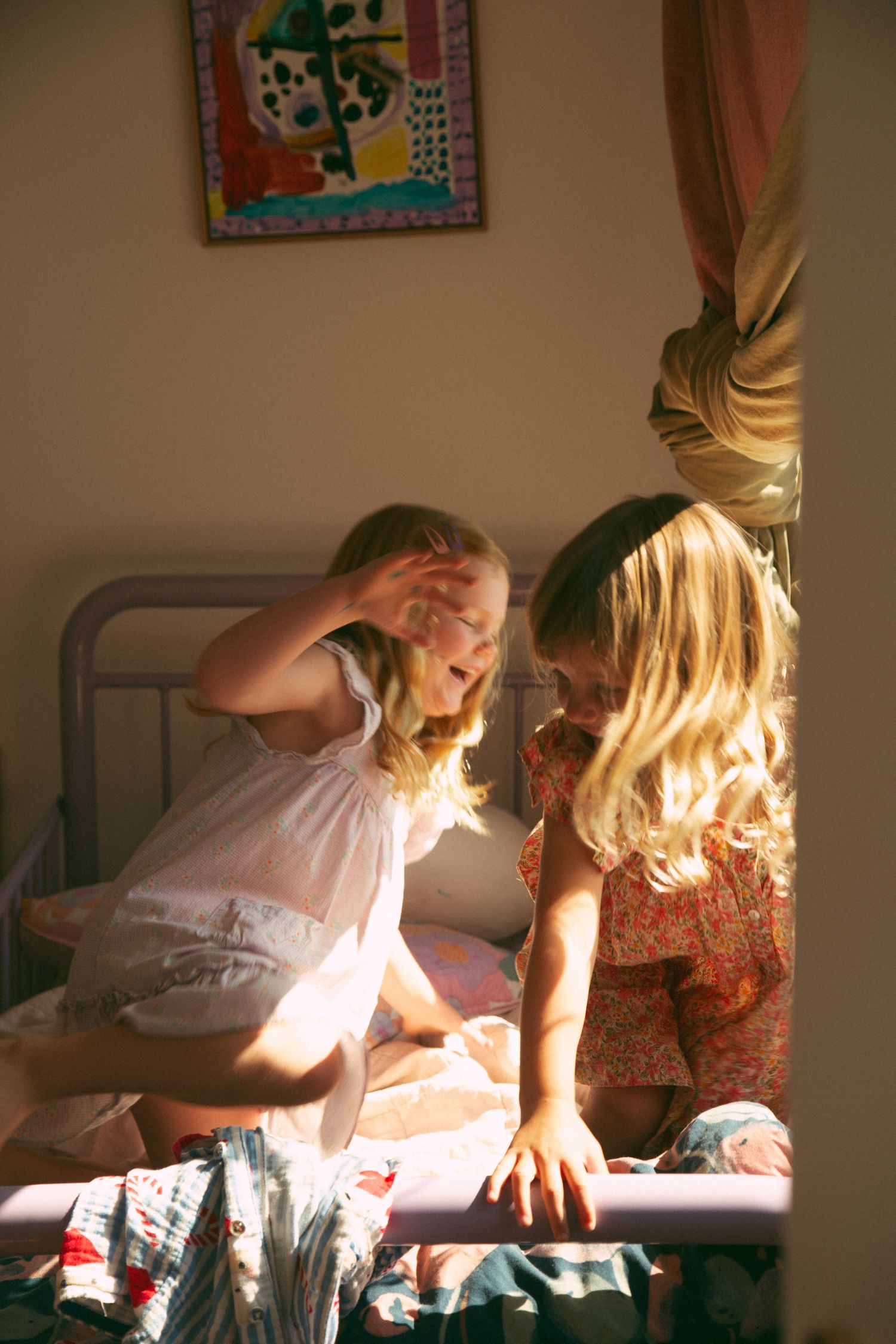 Two children share a playful moment together on a bed in a sunlit room with a framed artwork on the wall.