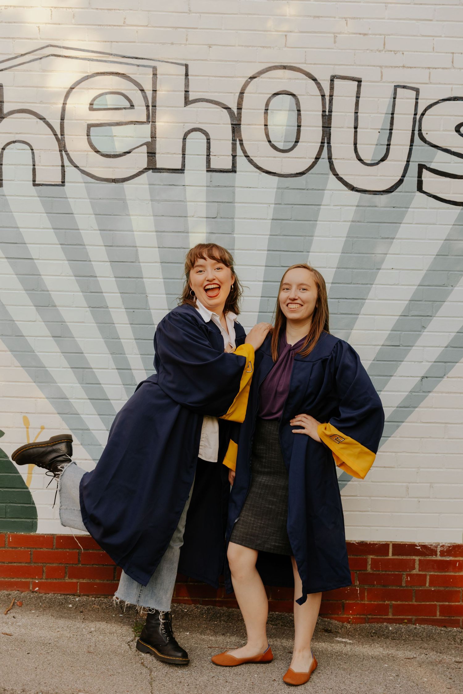 Two people in blue graduation gowns pose together in front of a stylized wall mural with radiating lines.