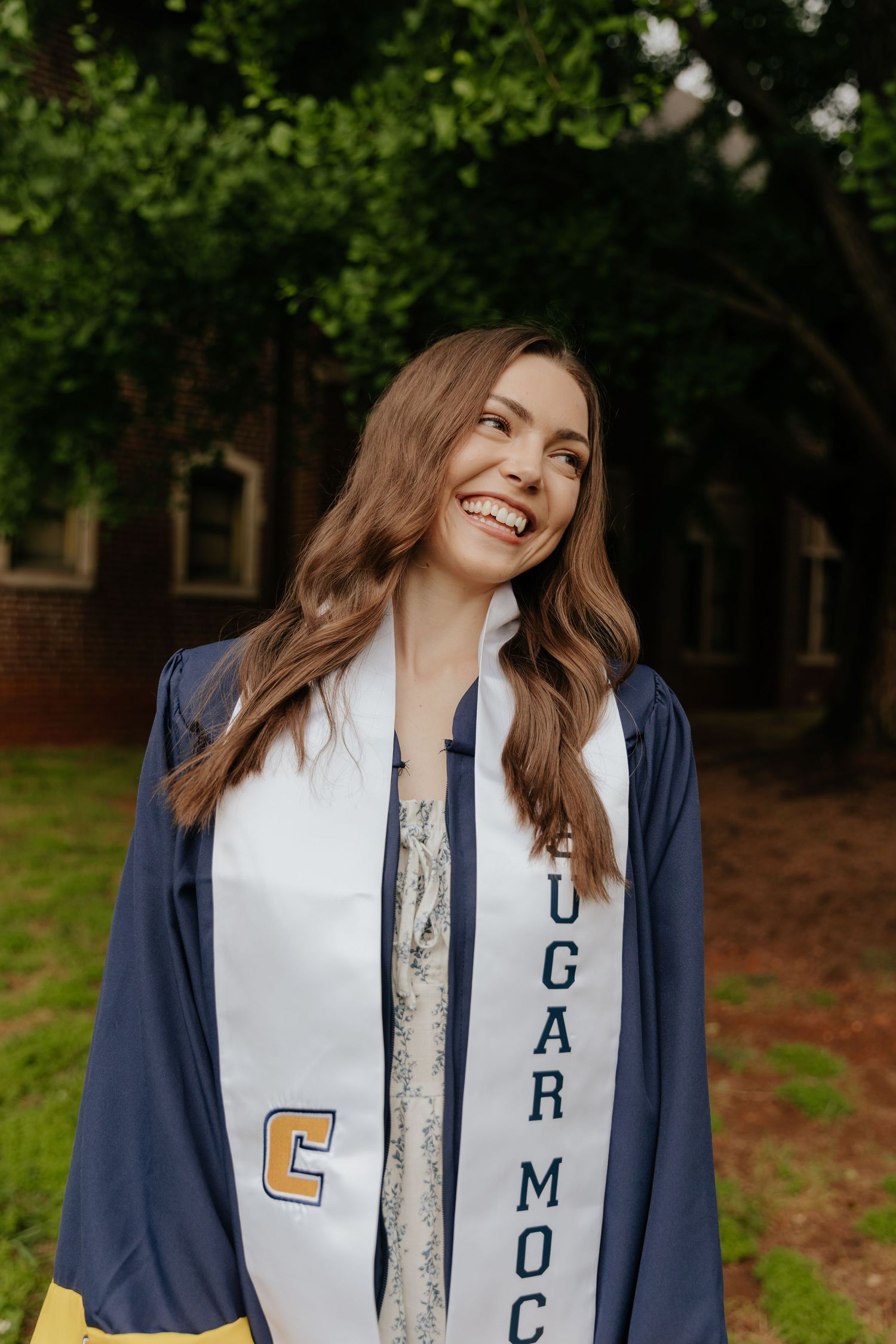 Graduate in navy blue cap and gown wearing white sash smiles joyfully during outdoor graduation photoshoot.