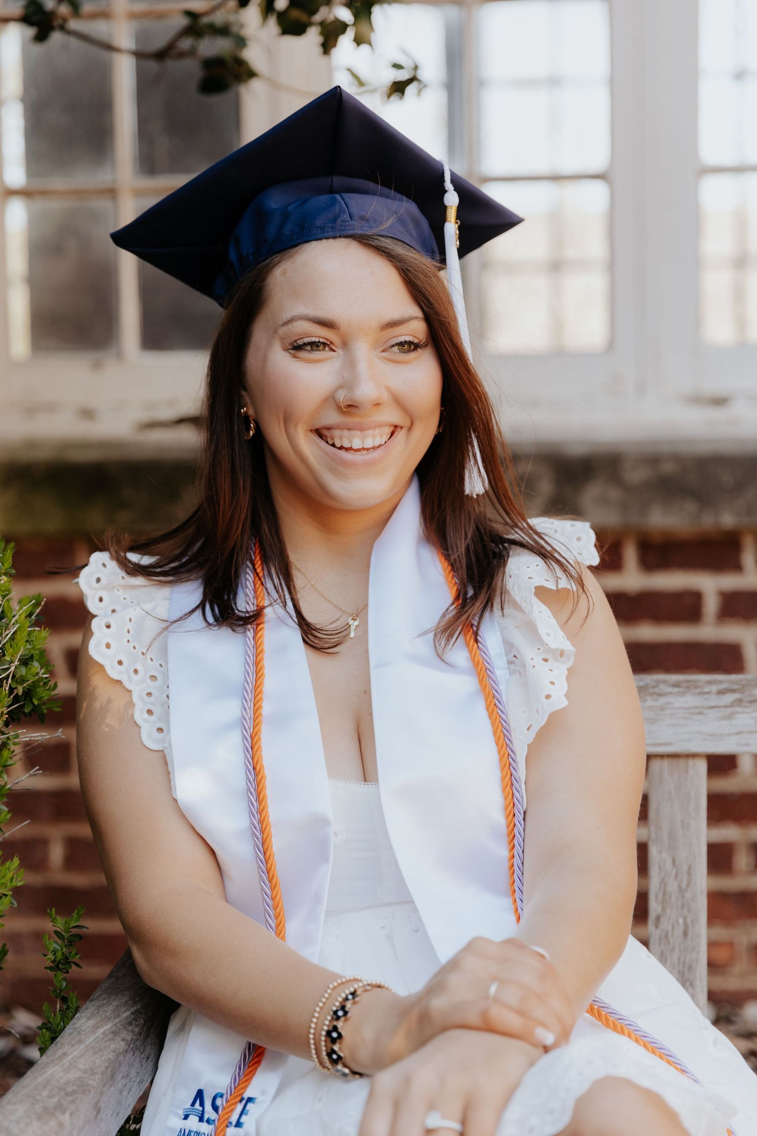 A graduate in a white dress and cap poses outdoors on a bench with a bright smile and orange stole.