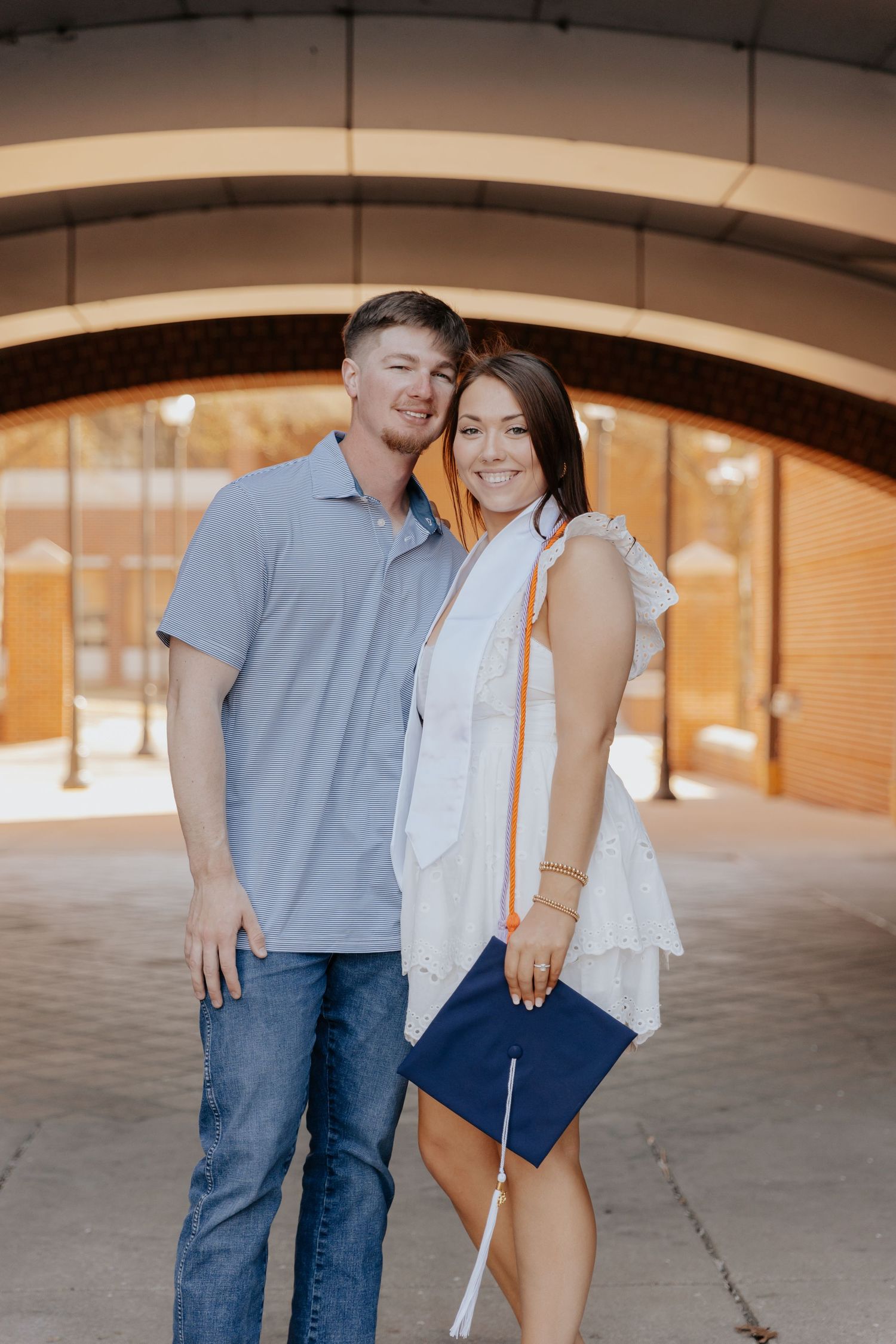 A couple poses for a graduation portrait under an archway with warm lighting in the background.