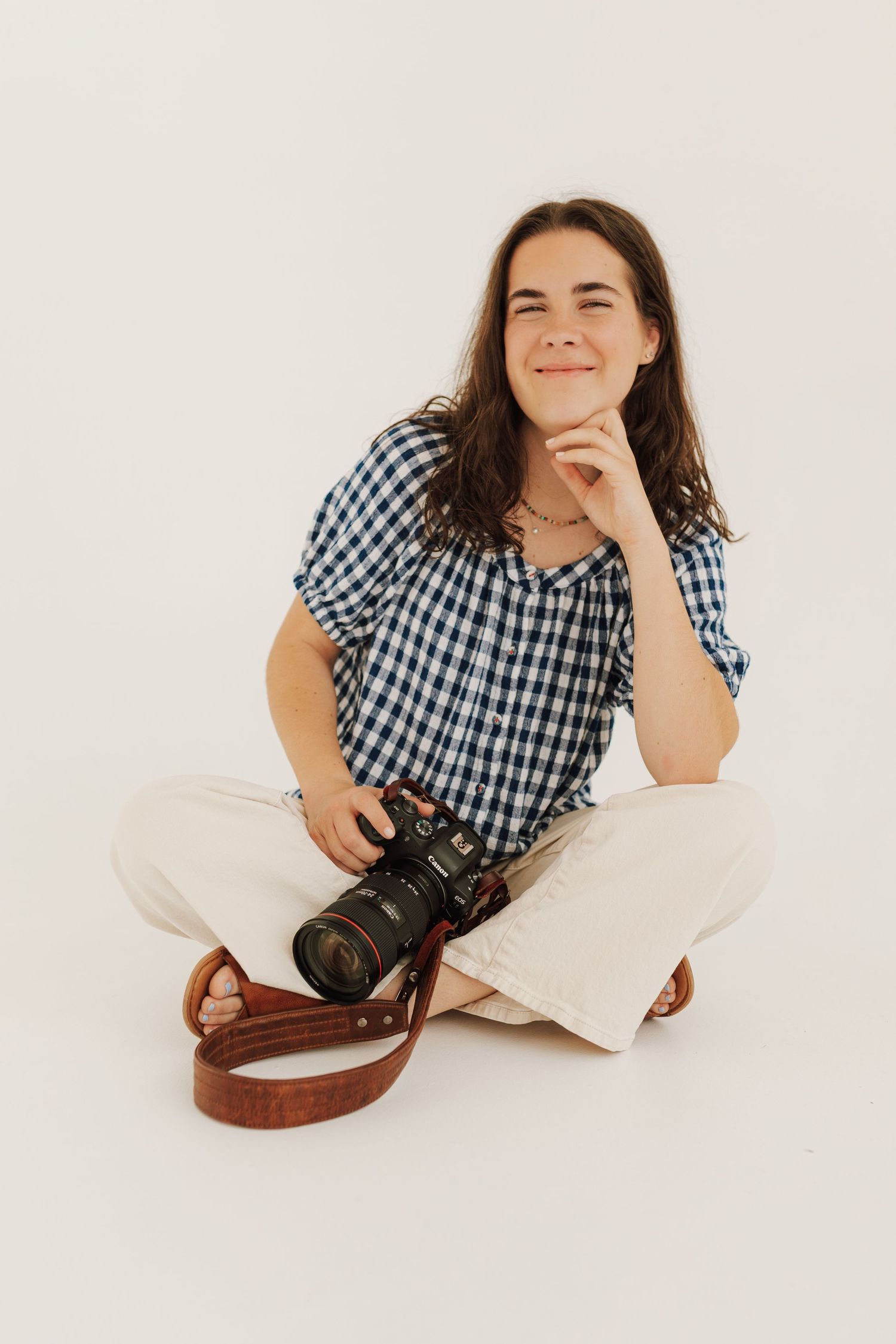 A photographer wearing a gingham shirt and white pants sits cross-legged on white background with a professional camera.