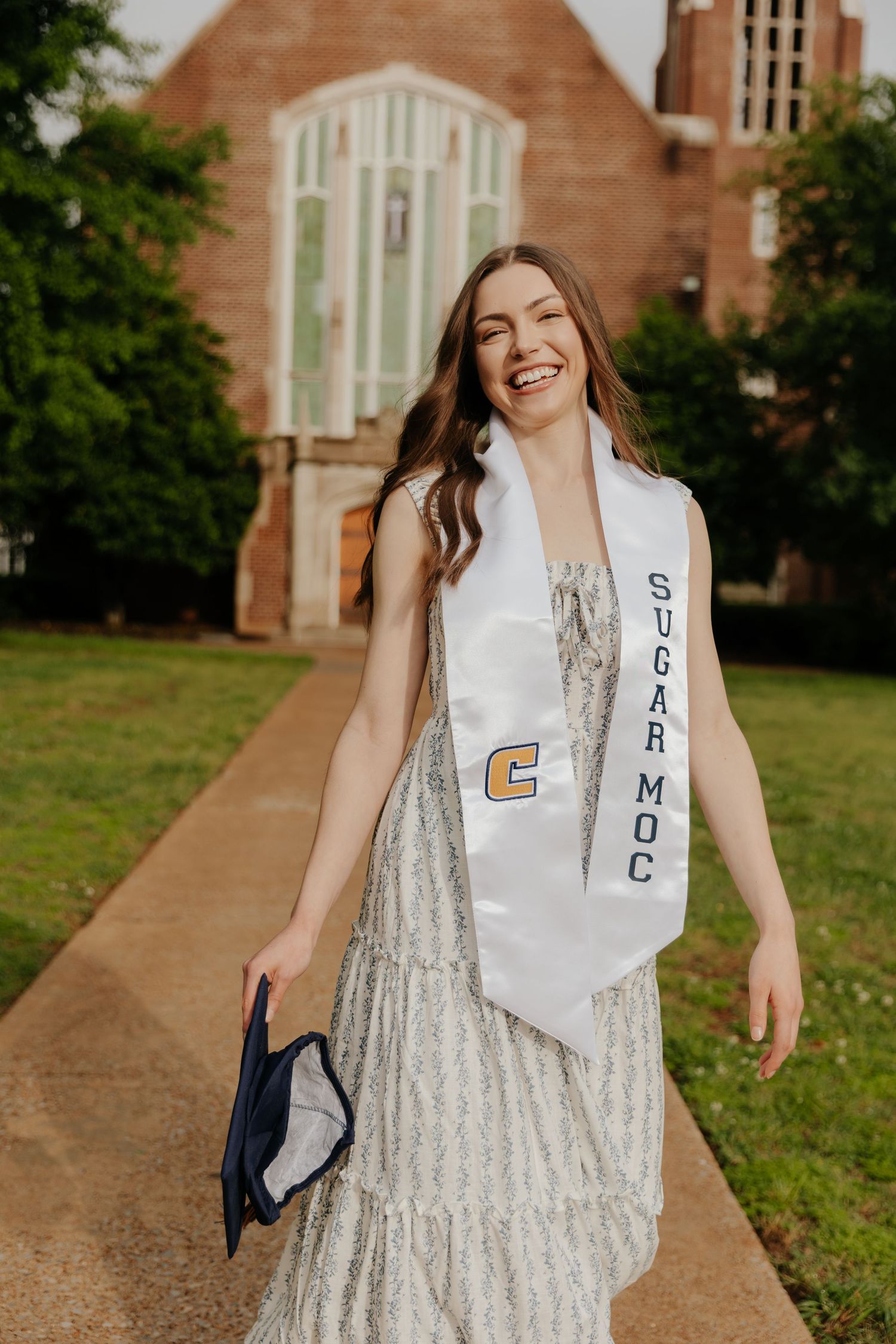 Graduate in white lace dress and graduation stole poses on campus walkway with brick church in background.