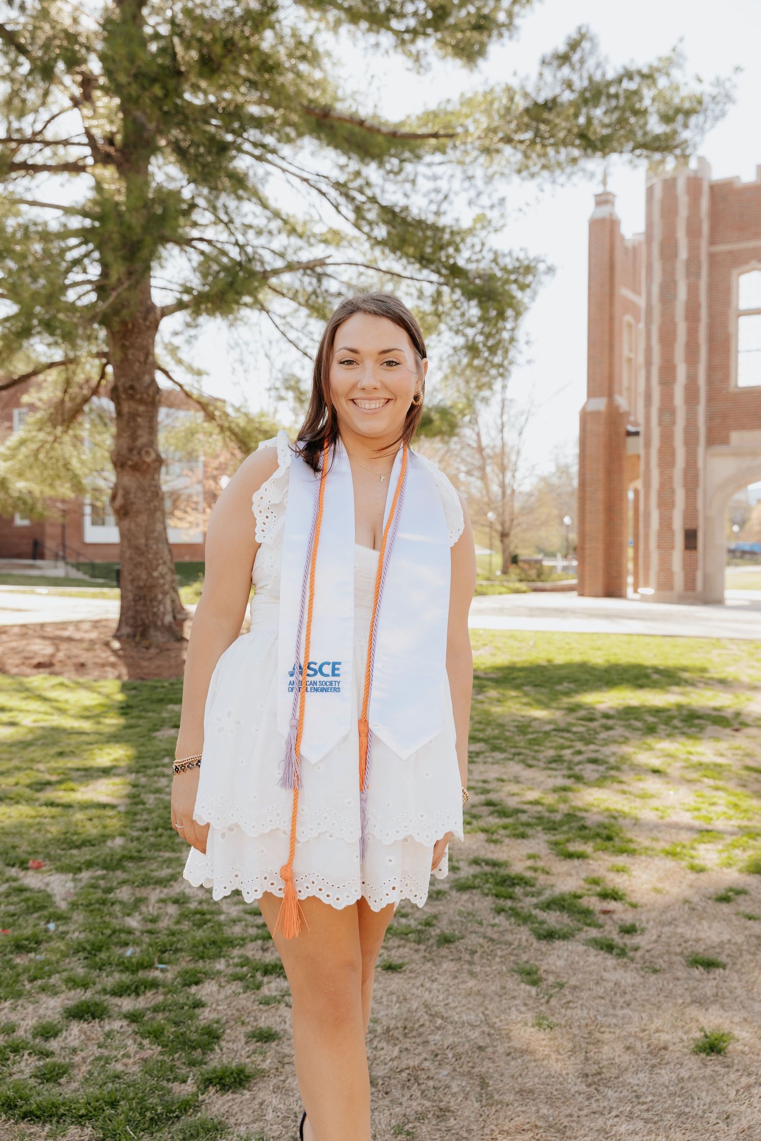 A graduate stands outdoors in a white lace dress with orange graduation stoles on a sunny spring day near campus buildings.
