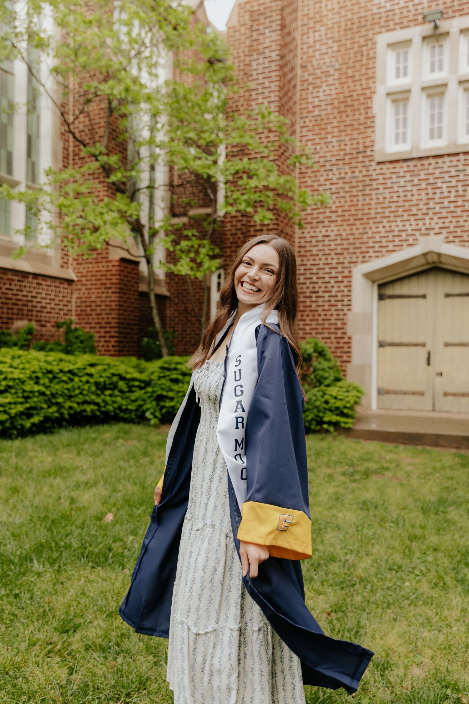Graduate in lace dress and blue graduation gown poses in front of brick university building with yellow pouch.