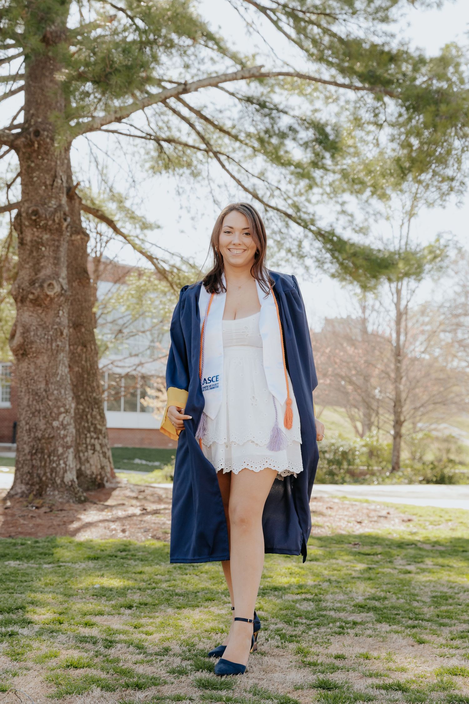 A person wearing a white dress and navy blue coat poses outdoors under a pine tree on a sunny spring day.