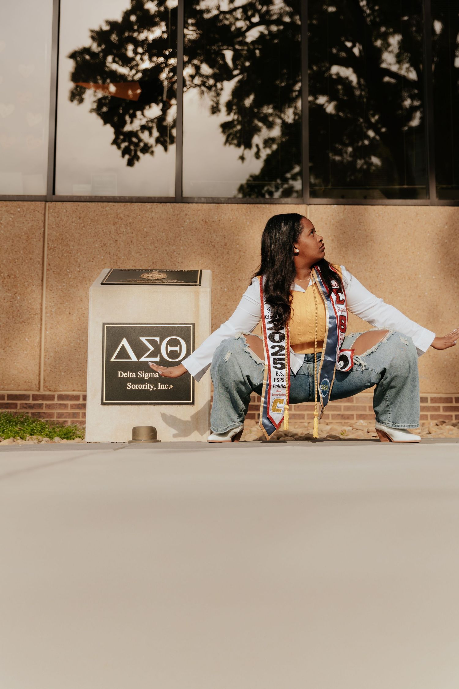 A student crouches beside a Delta Sigma Theta sorority sign wearing a yellow top, white jacket and decorated sash.