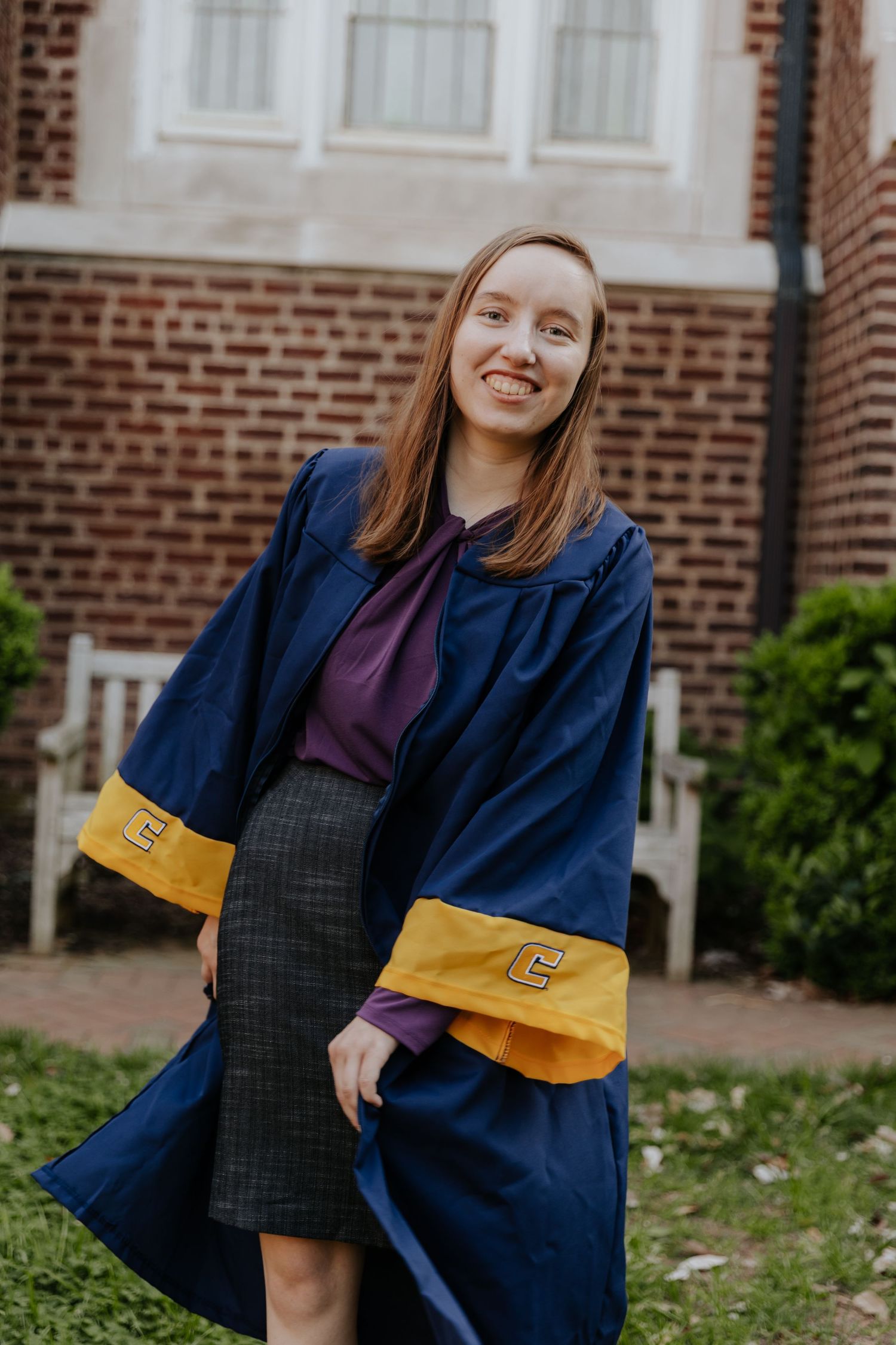 Graduate wearing blue and gold academic regalia with a purple top and pencil skirt stands outside a brick building.