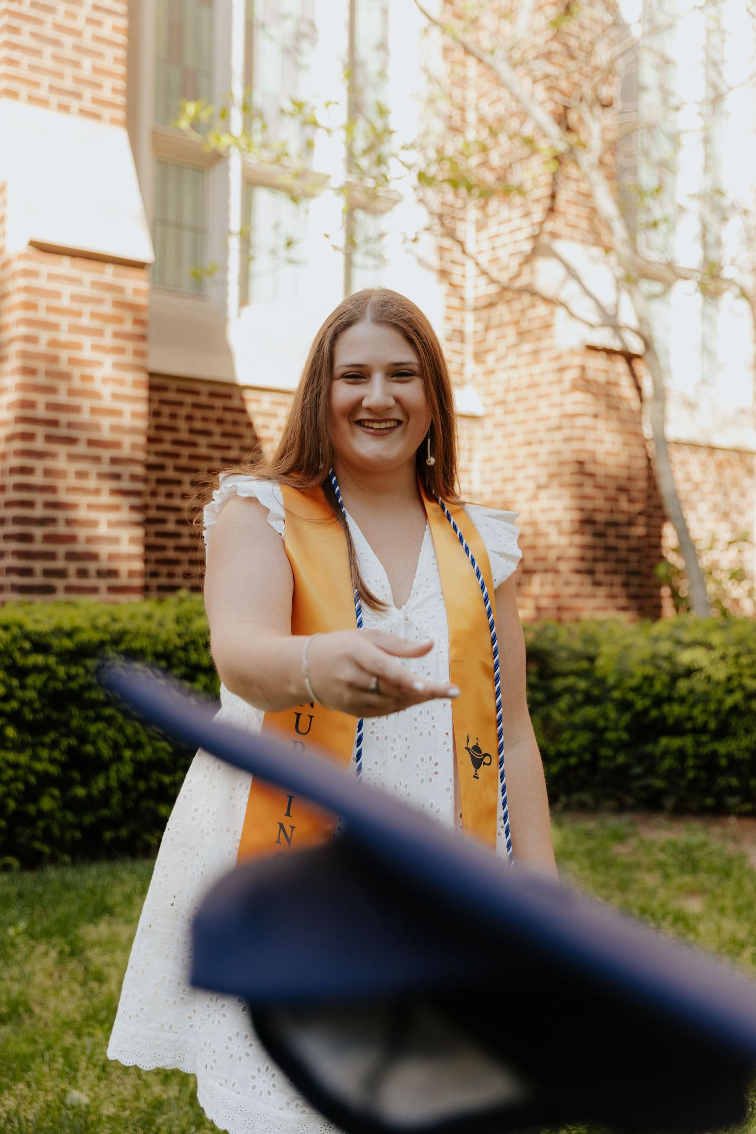 Someone standing outside a brick building wearing a white eyelet dress with orange accents and a braided crossbody bag.