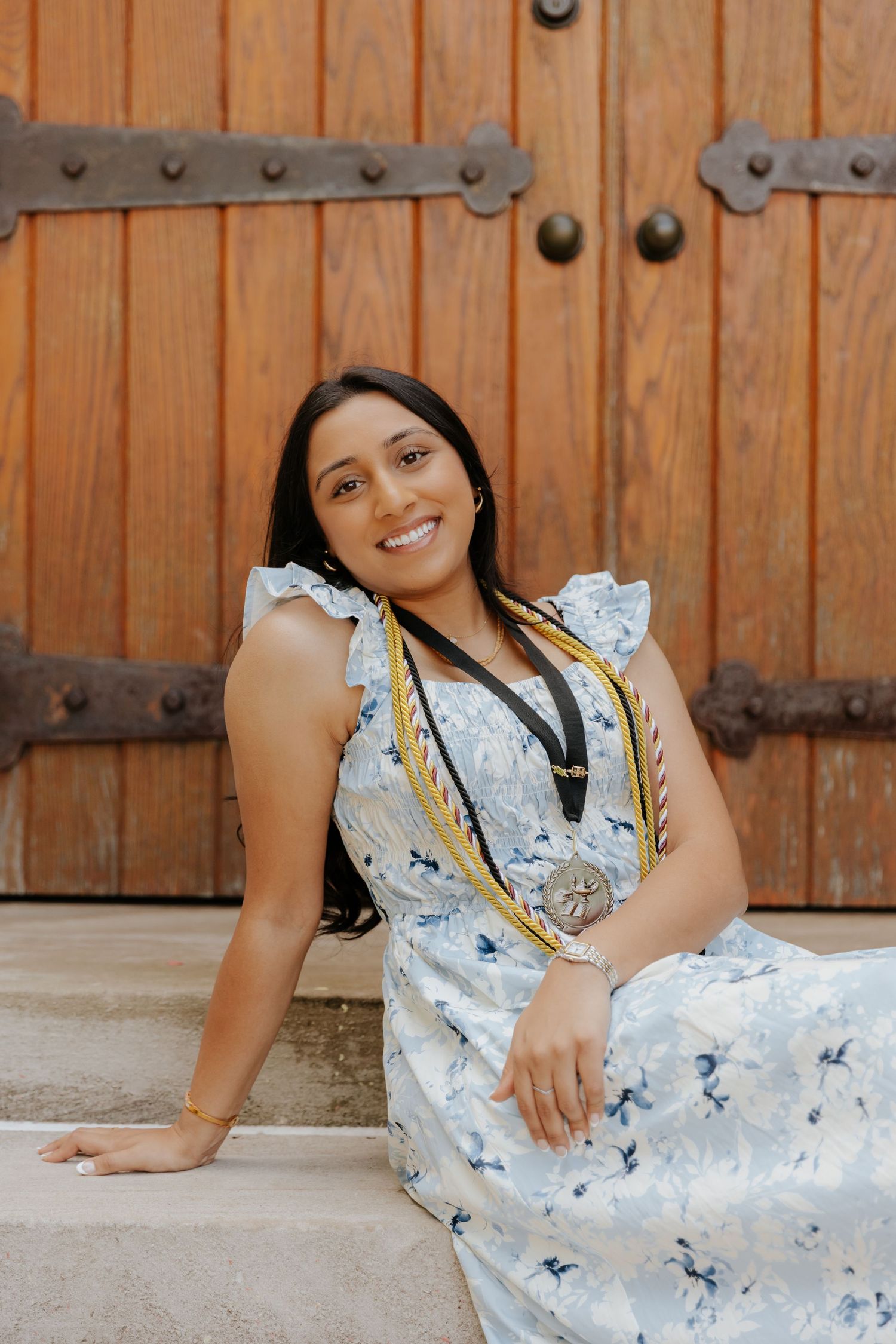 A woman in a light blue floral dress sits on stone steps in front of a rustic wooden door with metal hardware.