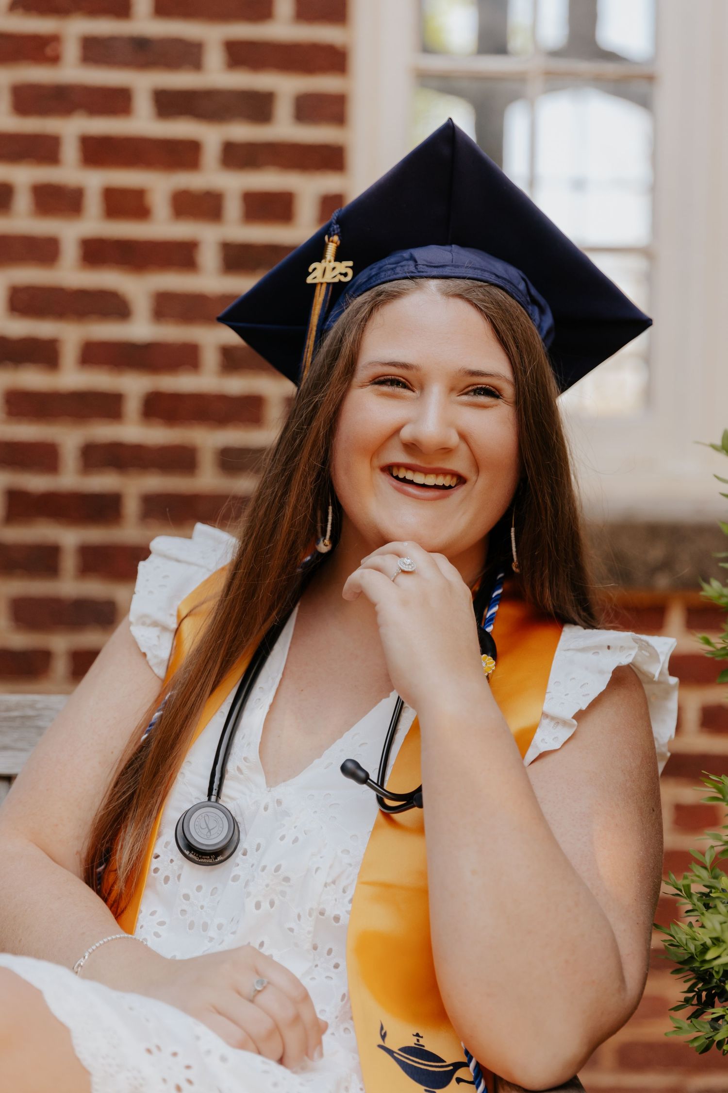 A graduate wearing a cap with medical stethoscope and white eyelet dress smiles joyfully against a brick wall backdrop.
