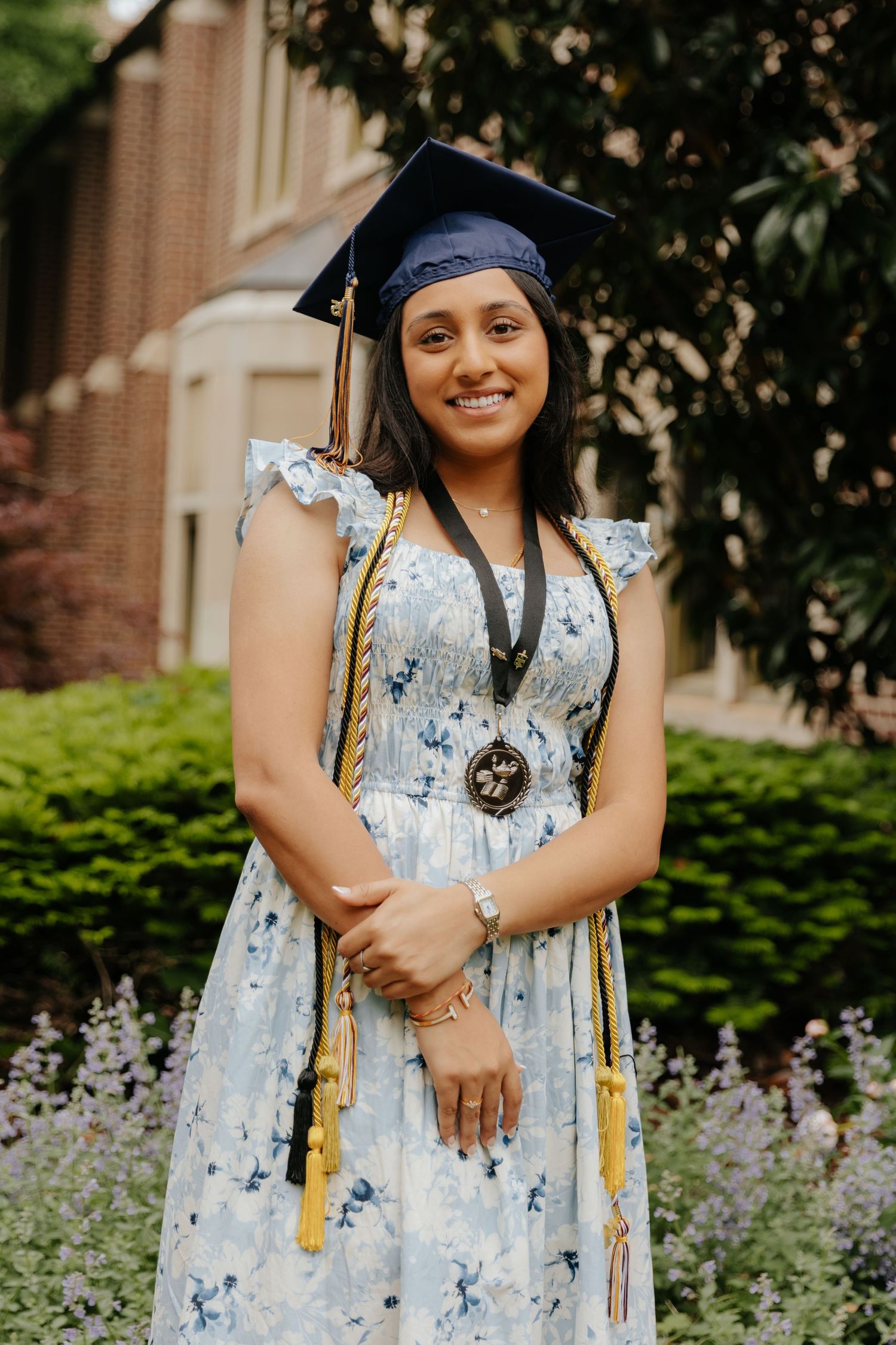 Graduate in blue cap and gown wears floral dress with honor cords while posing outdoors by brick building.