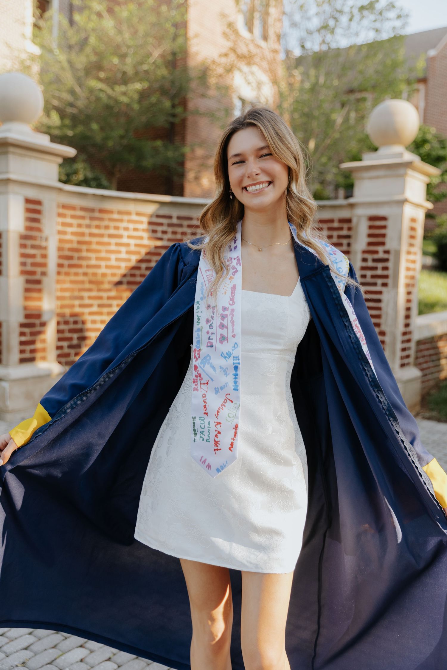 A person in a navy coat shows off their white dress with floral scarf on a brick courtyard.