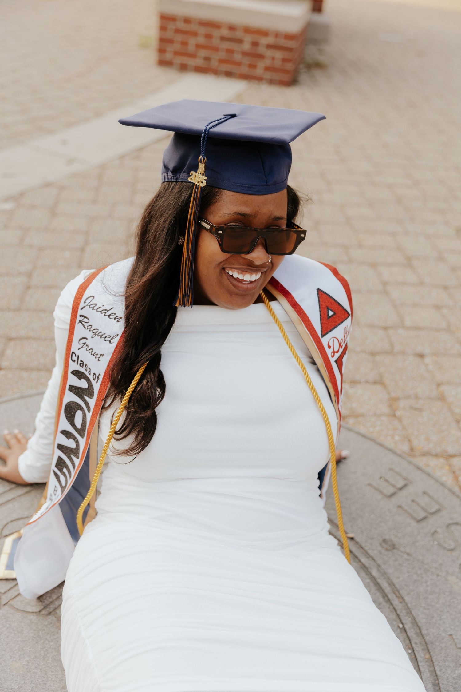 Graduate in white dress and cap smiling while sitting outdoors wearing sunglasses and graduation stole.