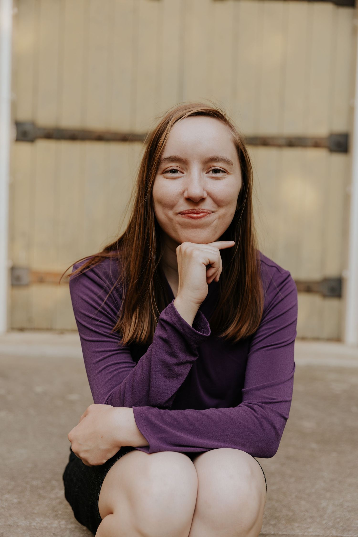 A person in a purple sweater sits outdoors with chin resting on hand while smiling warmly at the camera.
