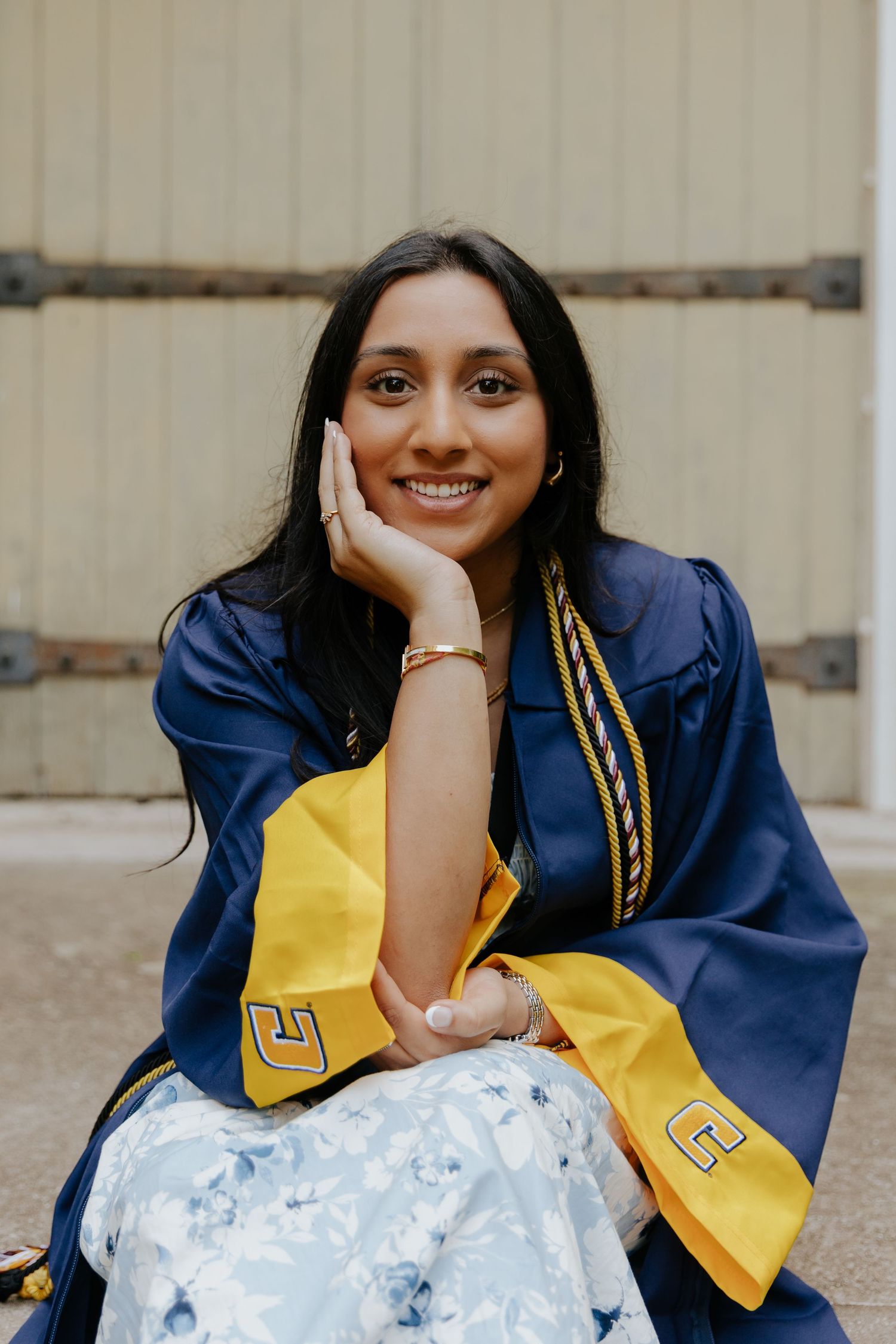 Graduate in blue and gold academic regalia sits casually on steps with hand on face, wearing floral skirt.