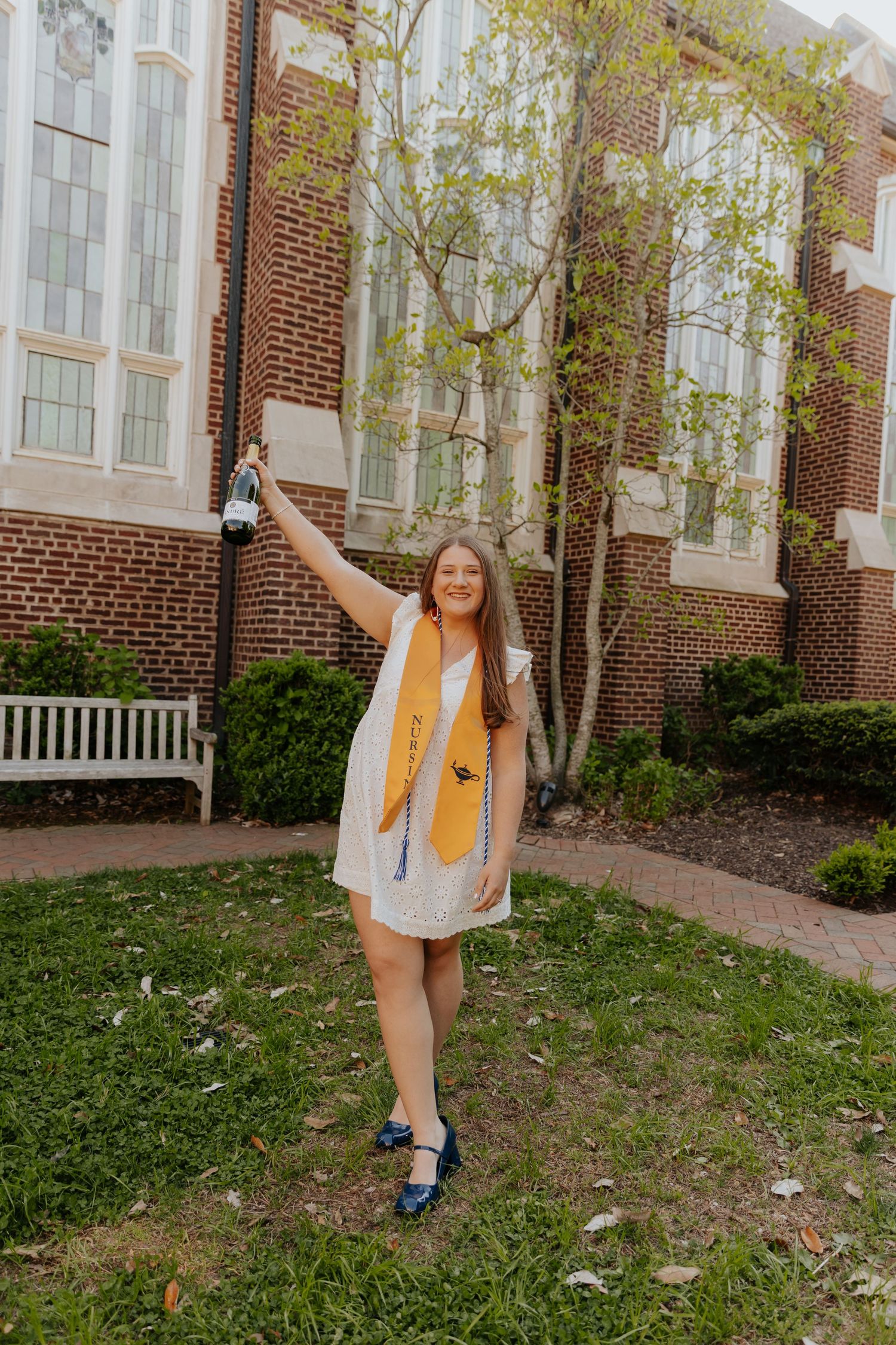 A joyful graduate in a white dress and yellow scarf celebrates outside a brick university building in springtime.
