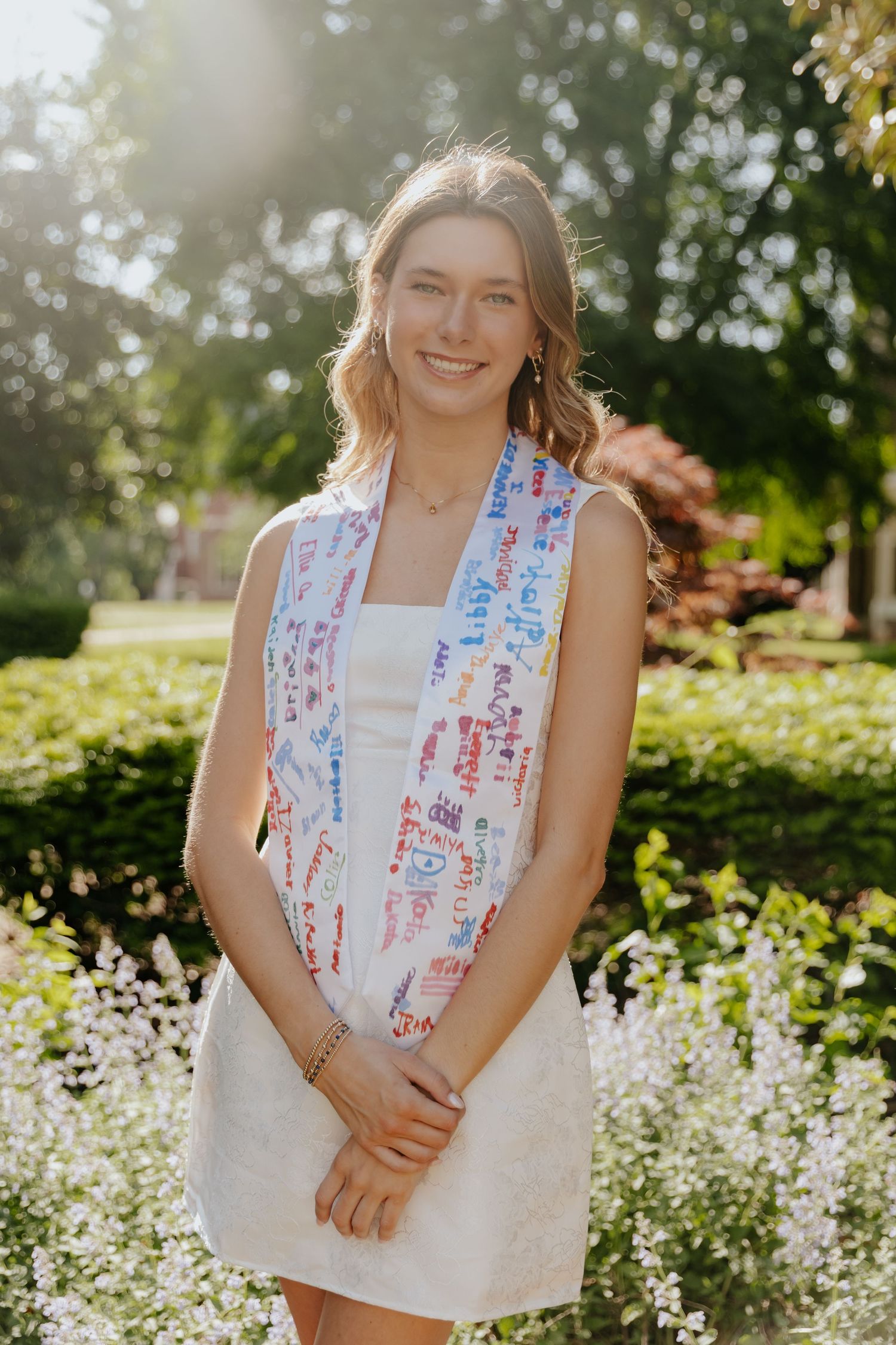 A person in a white dress and patriotic red white and blue scarf smiles outdoors on a sunny summer day in a garden.