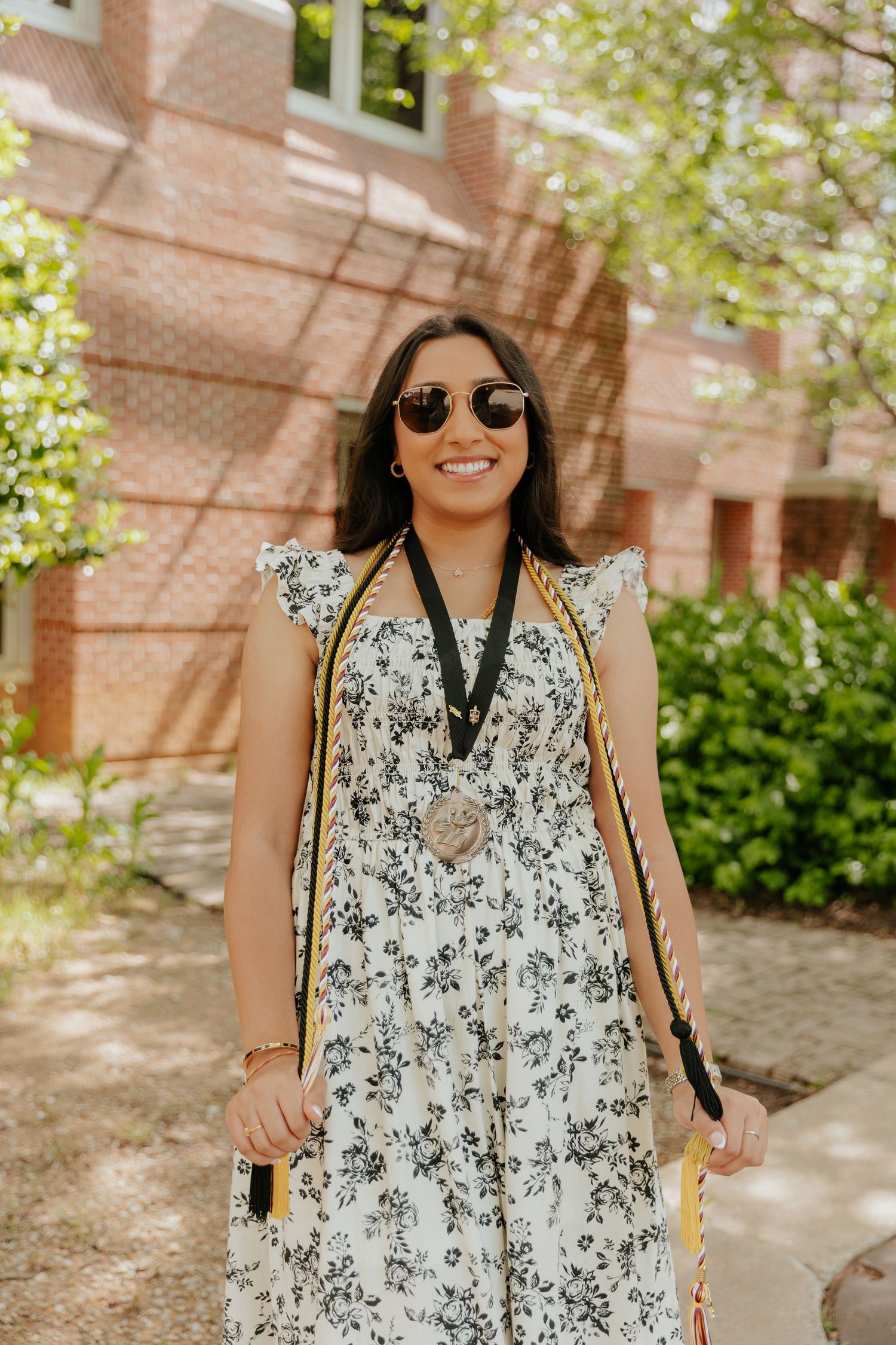 Someone wearing a floral print summer dress with sunglasses smiles on a sunny day near a brick building.